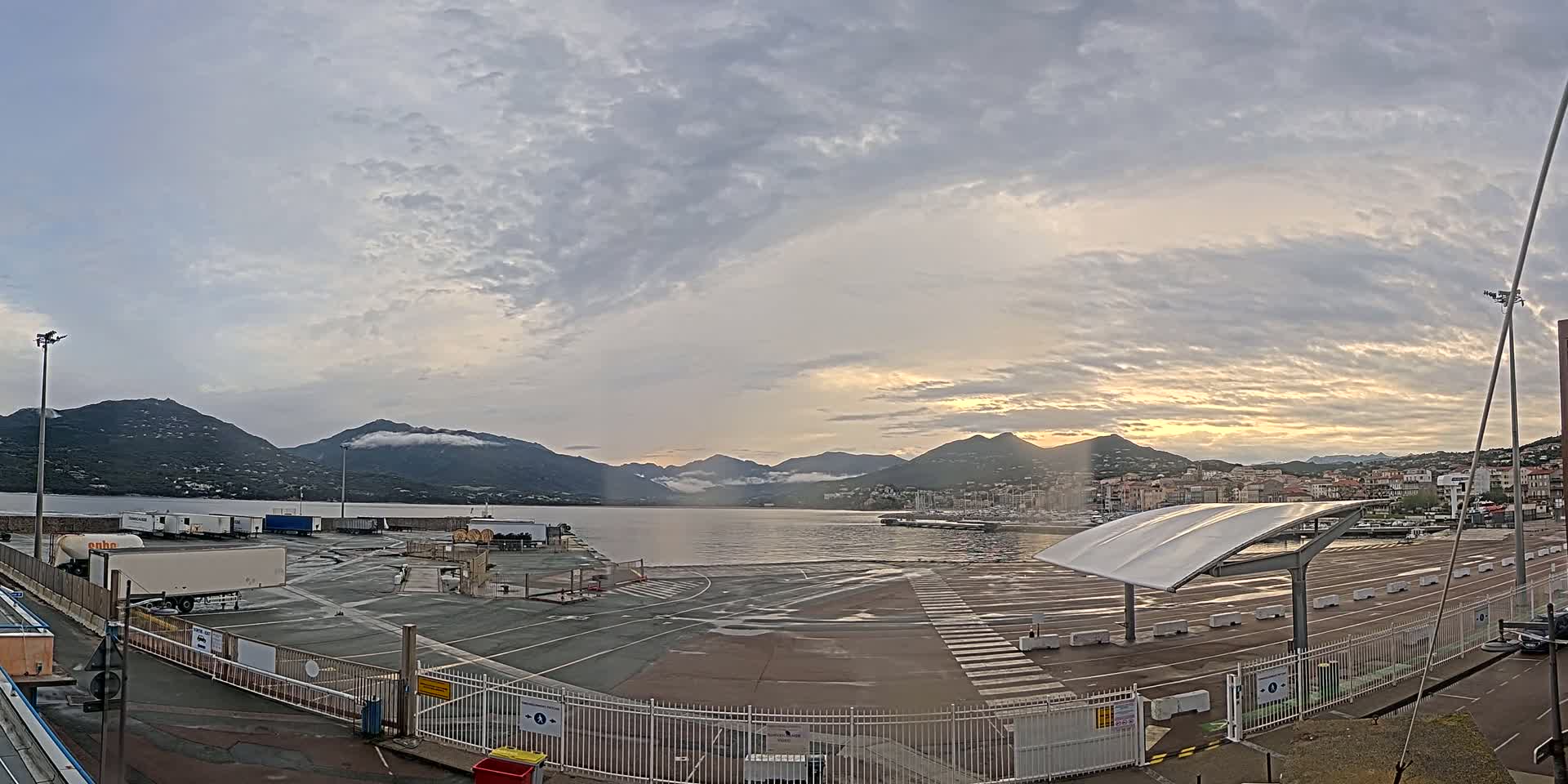 A mostly cloudy sunrise illuminates a harbor with several docked boats and a large empty paved area with semi-trucks parked near a fence.