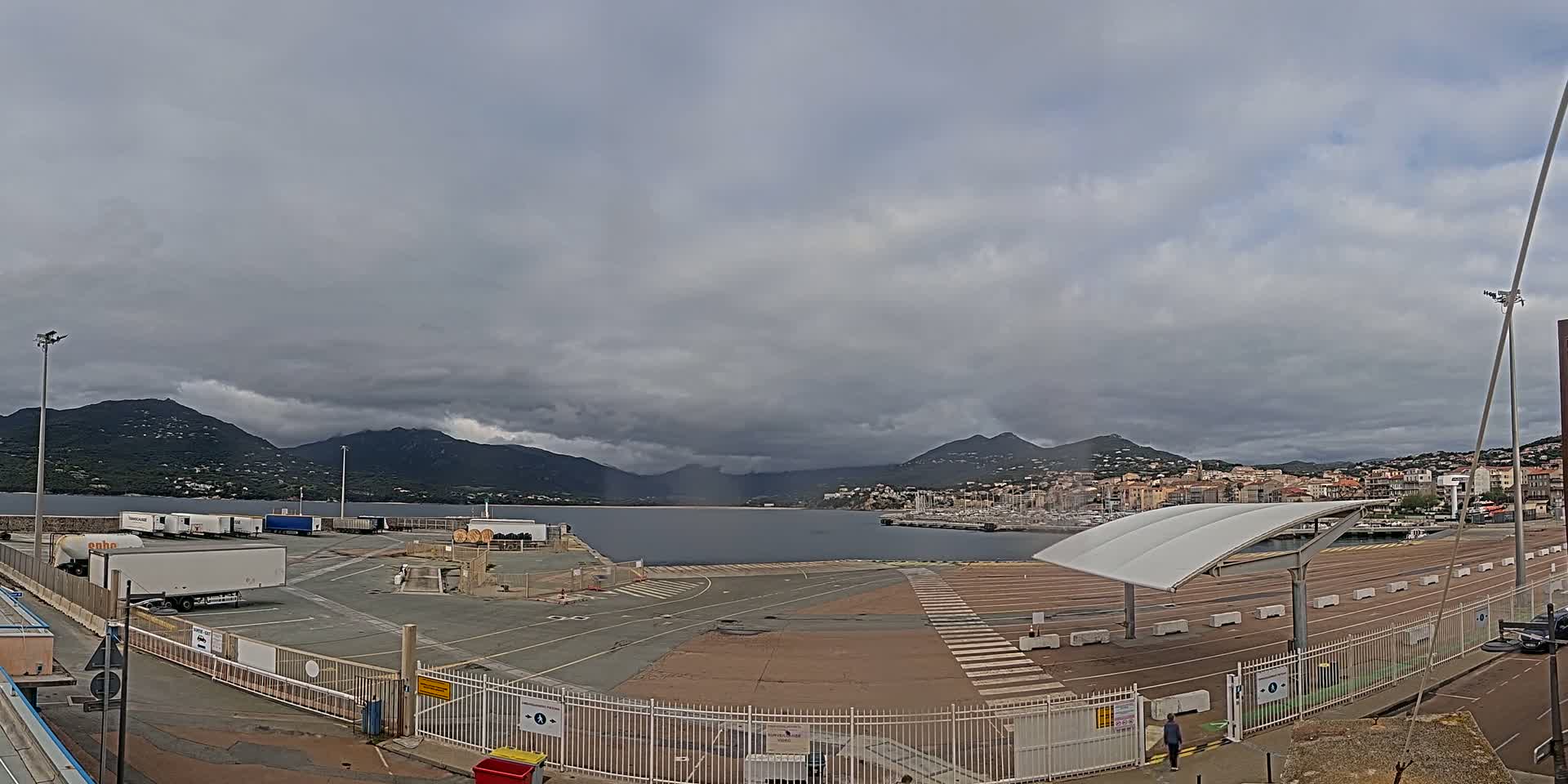 A large, empty port area with several parked trucks is seen under a cloudy sky, overlooking a calm body of water with a town and mountains in the background.