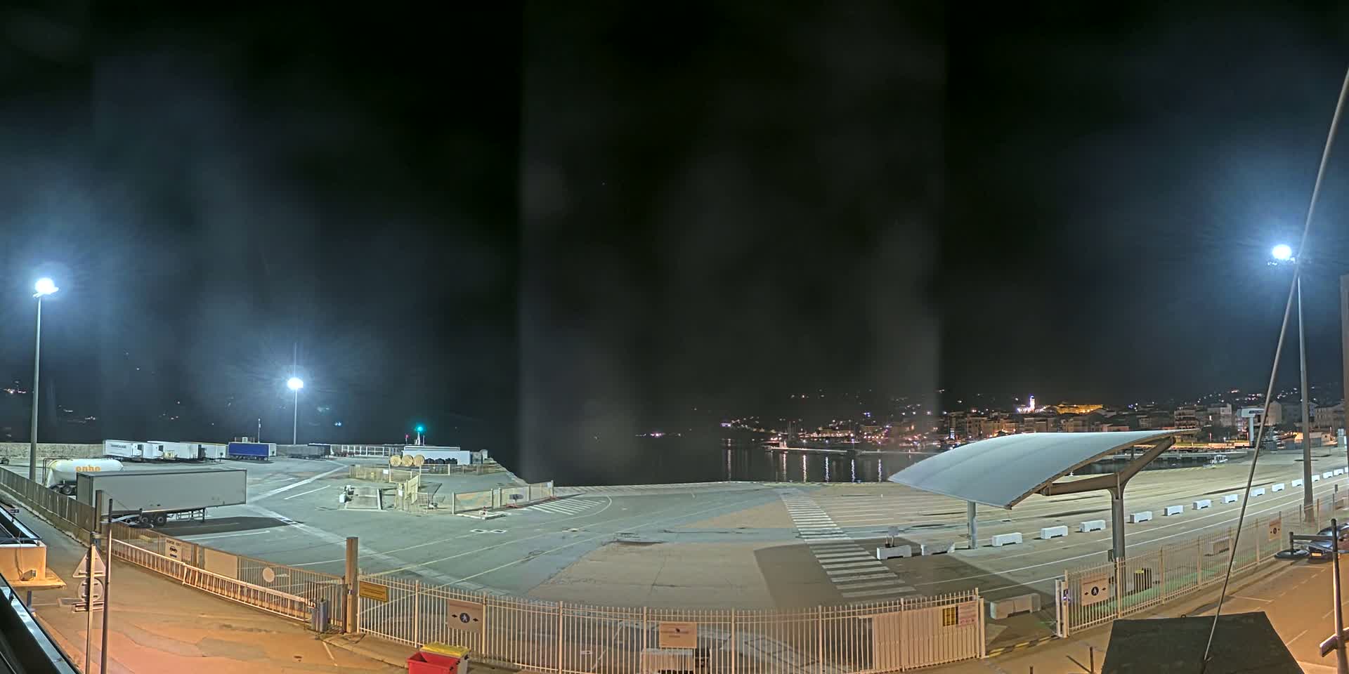 A nighttime view of a mostly empty port area with several trucks parked, under hazy, dark skies.