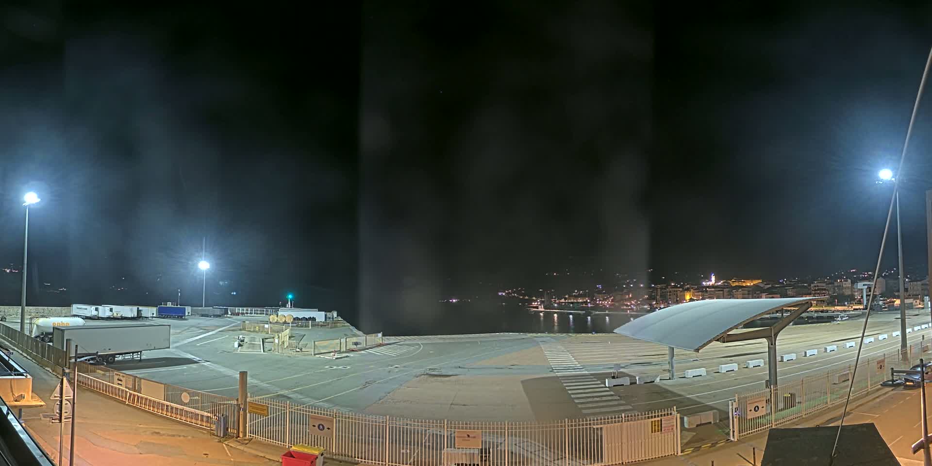 A nighttime view of an empty port area with several trucks parked near a body of water, under hazy conditions with distant city lights visible.