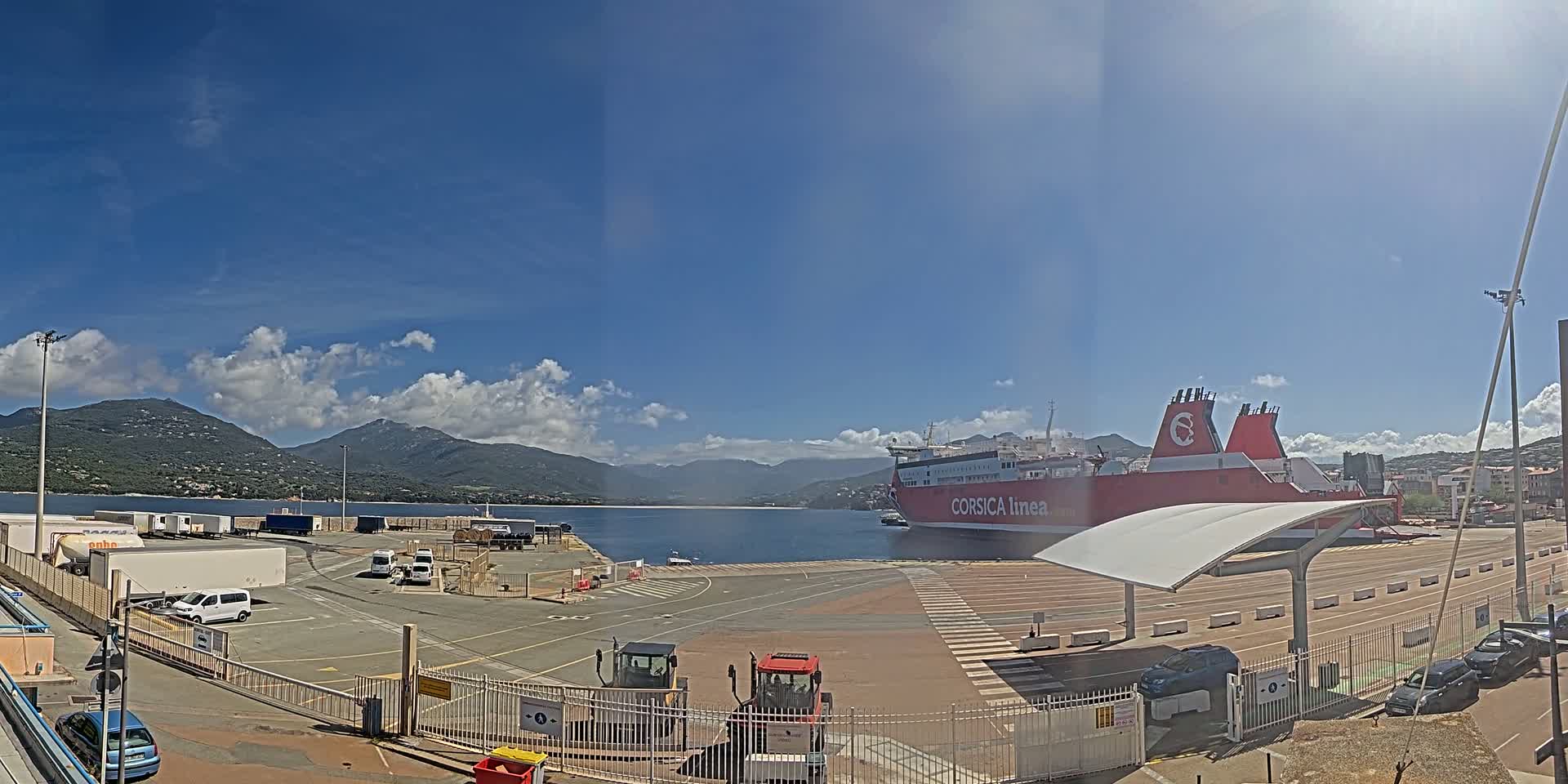 A large red and white ferry is docked at a port, under a mostly sunny sky with some scattered clouds, mountains are visible in the background.
