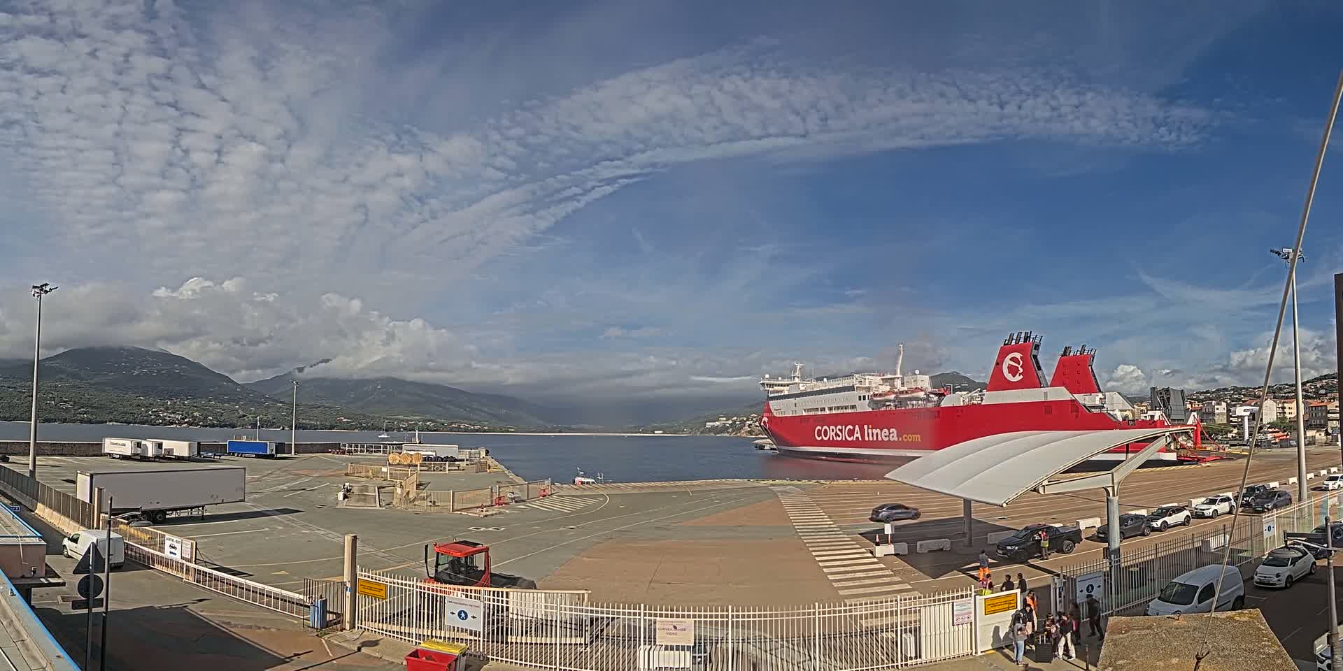 A large red ferry is docked at a port under a partly cloudy sky, with mountains in the background and several vehicles parked nearby.