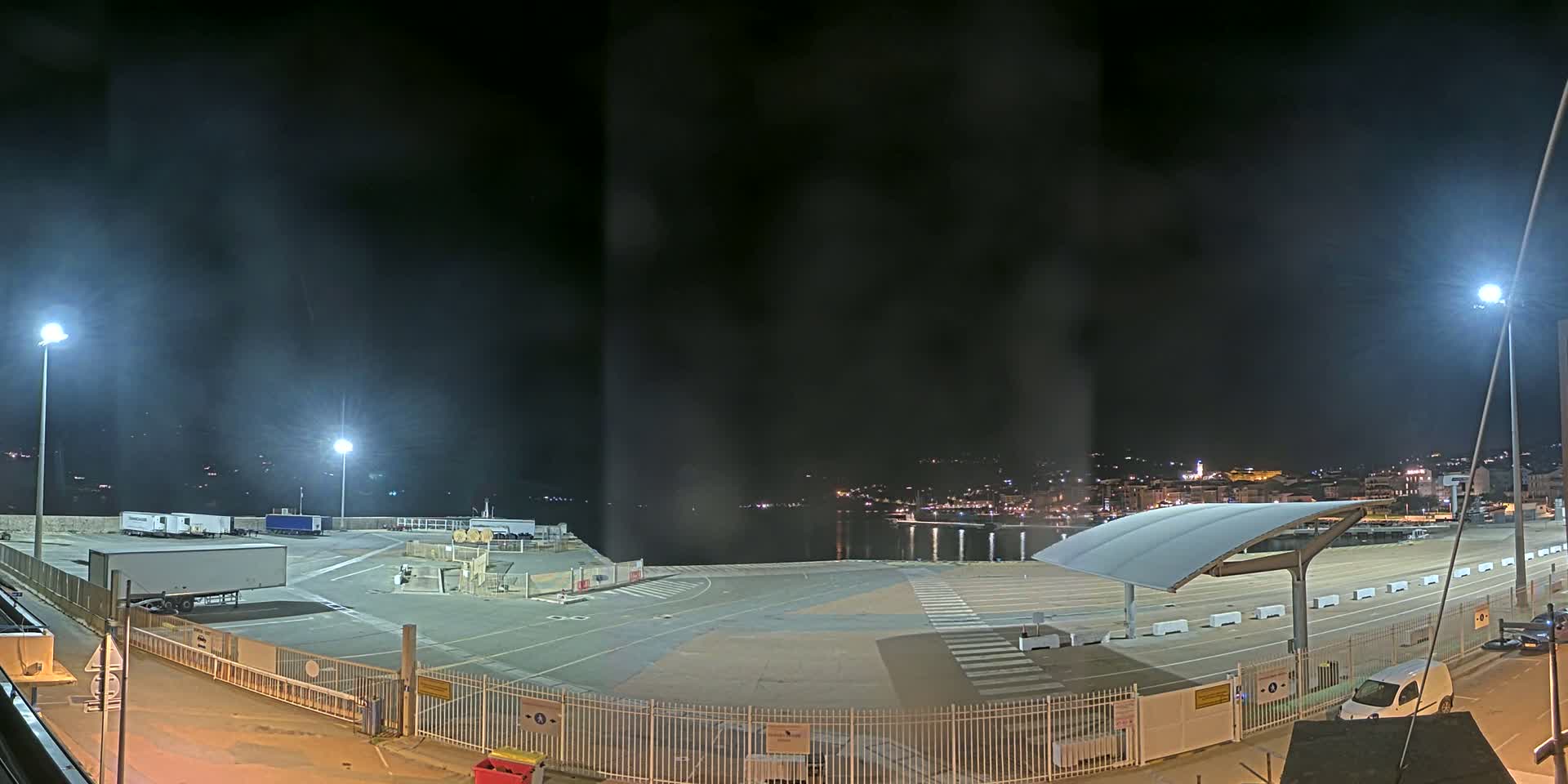 A nighttime view of an empty port area with several parked trucks and cars, illuminated by bright lights under a hazy, dark sky.