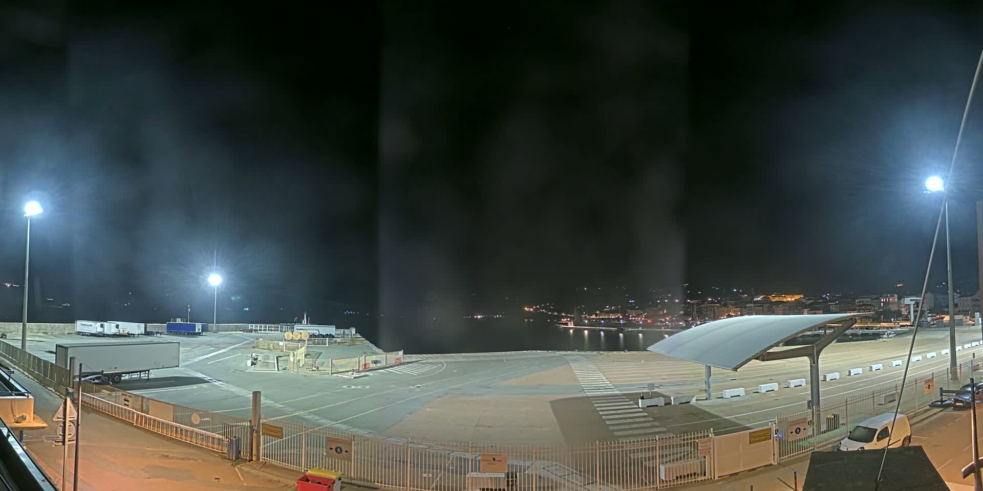 A nighttime view of a port area with several trucks and a covered waiting area, under hazy conditions.