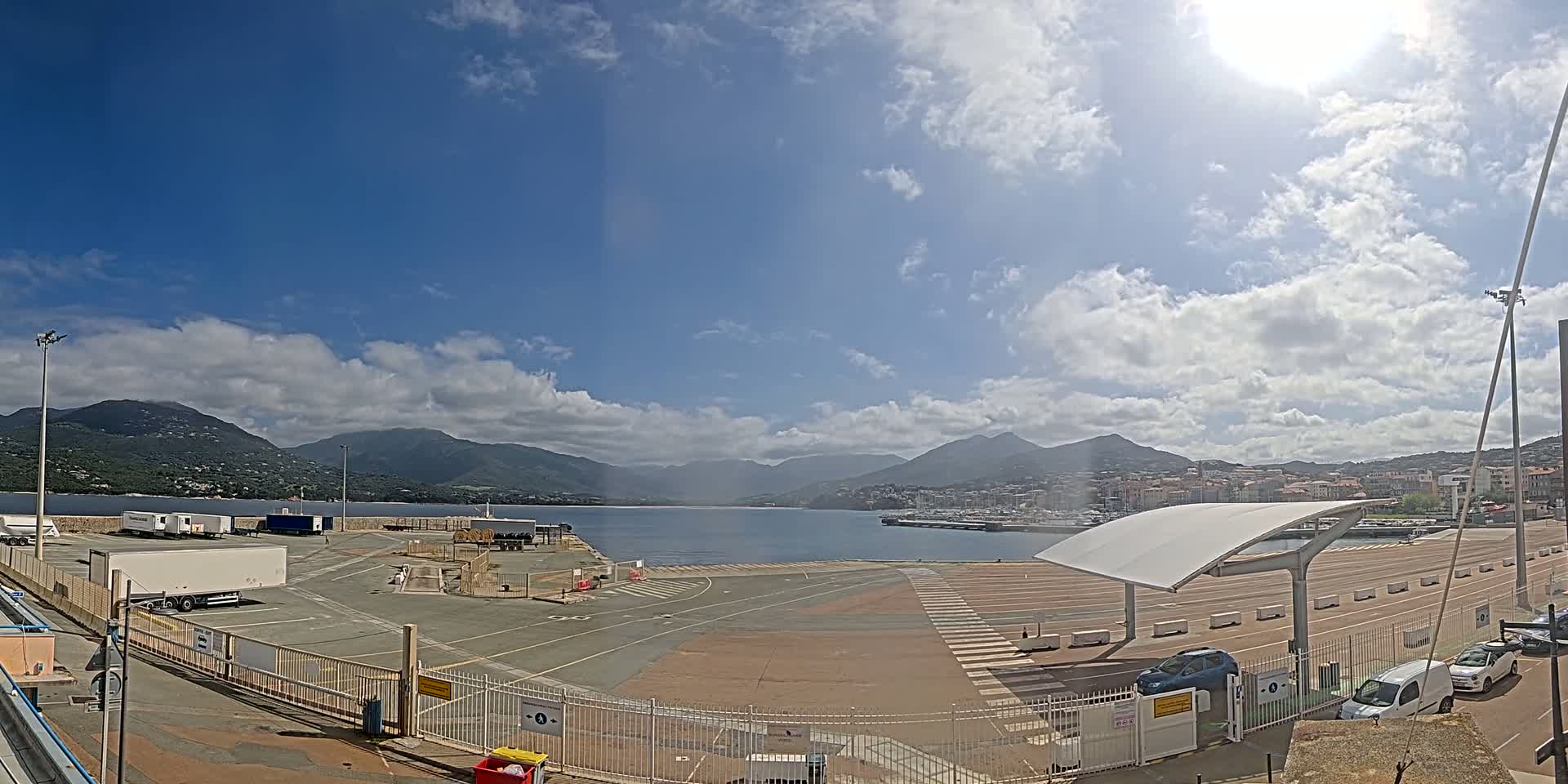 A sunny day with partly cloudy skies shows a port area with trucks, cars, and a large body of water with mountains in the background.