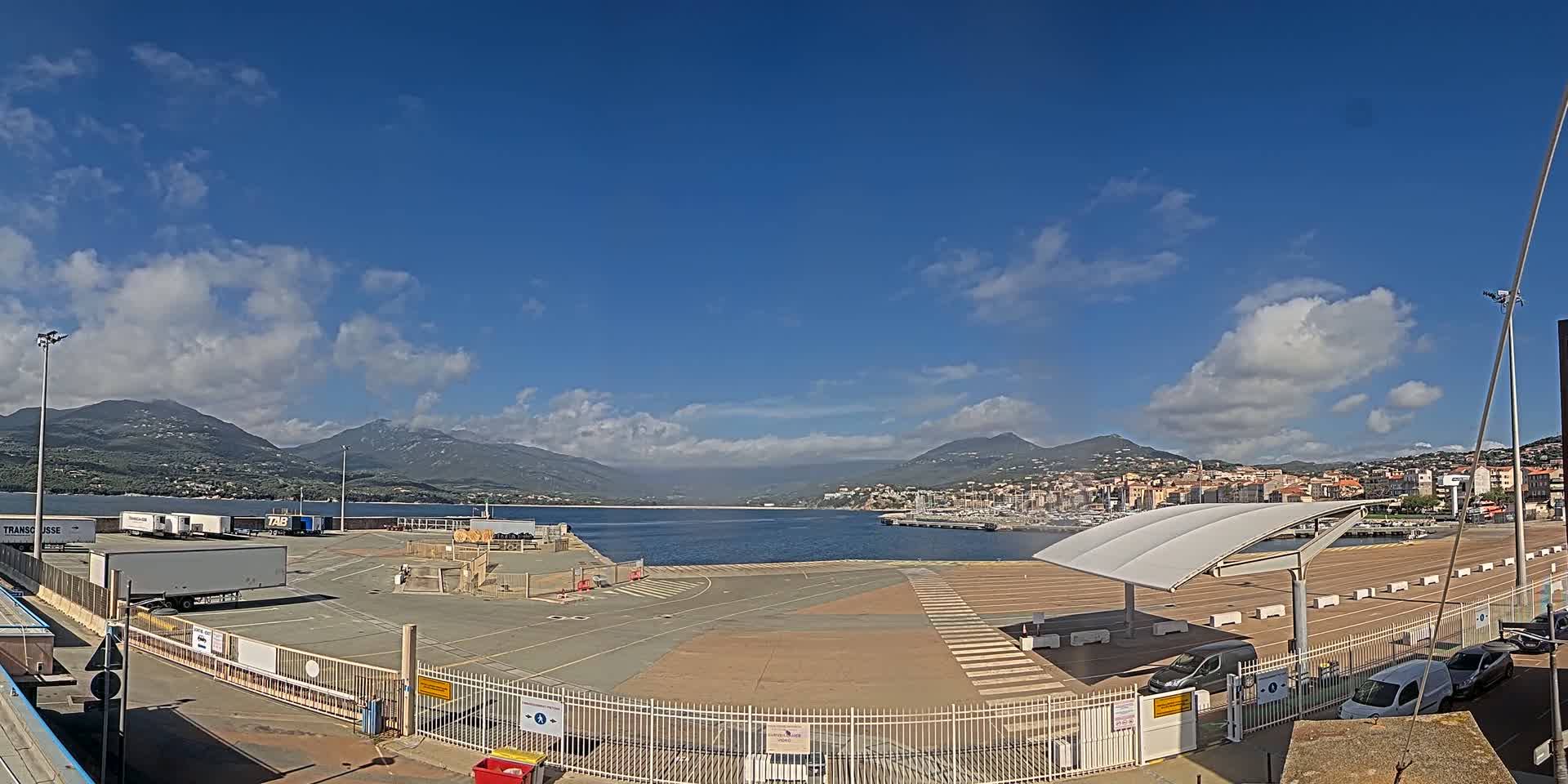 A mostly sunny day shows a port area with semi-trucks, parked cars, and a covered waiting area, all overlooking a calm body of water and mountains in the distance.