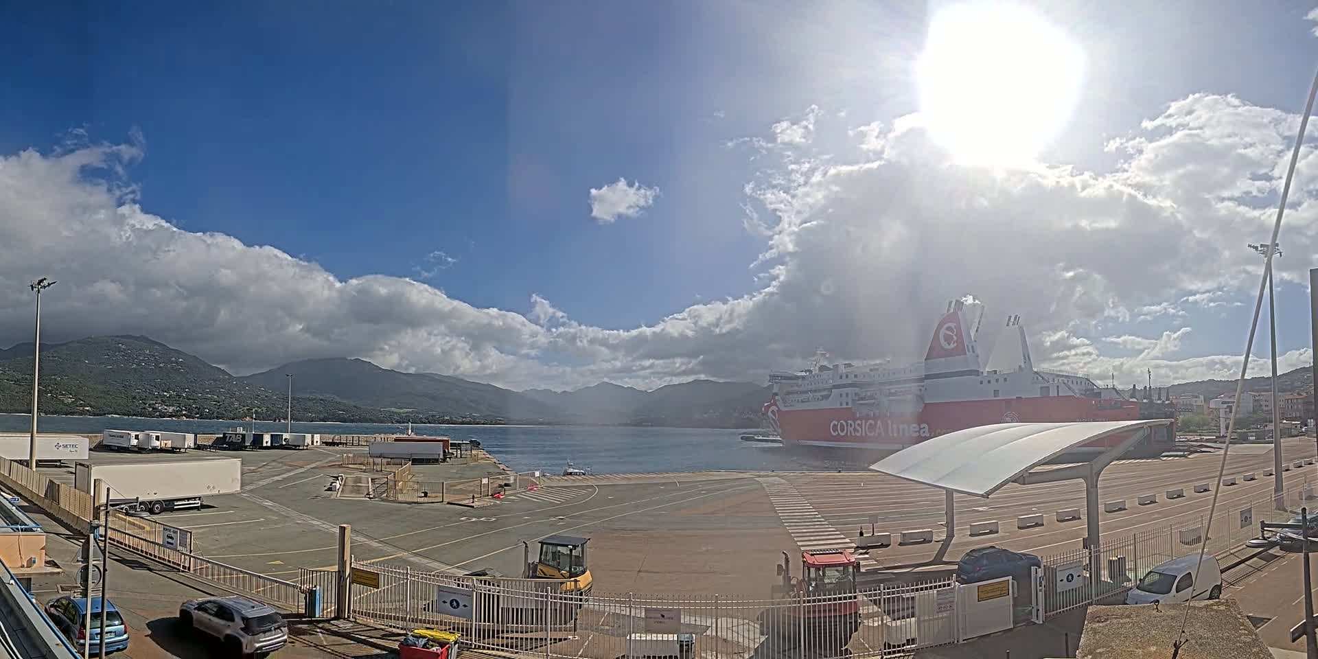 A large ferry is docked at a port under a partly sunny sky with fluffy white clouds, mountains are visible in the background.