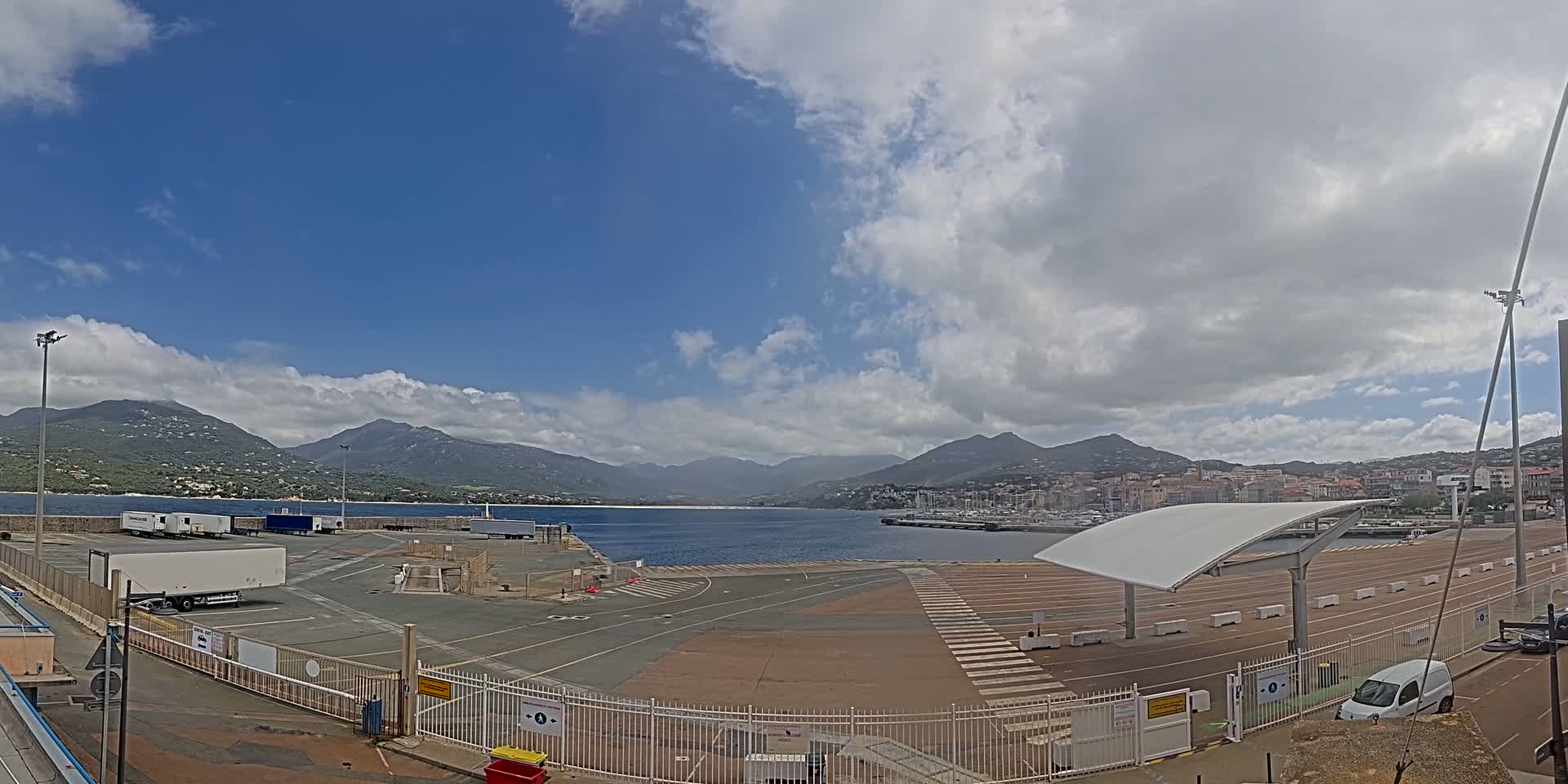 A mostly sunny day shows a port area with several parked trucks, a covered walkway, and a view of mountains and a town across the water.