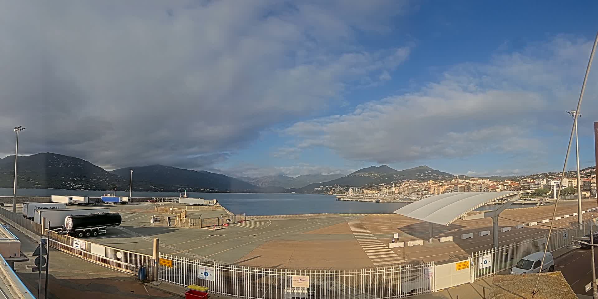 A partly cloudy day shows a harbor with several trucks and a large, empty paved area enclosed by a fence, with mountains and a town visible in the distance.