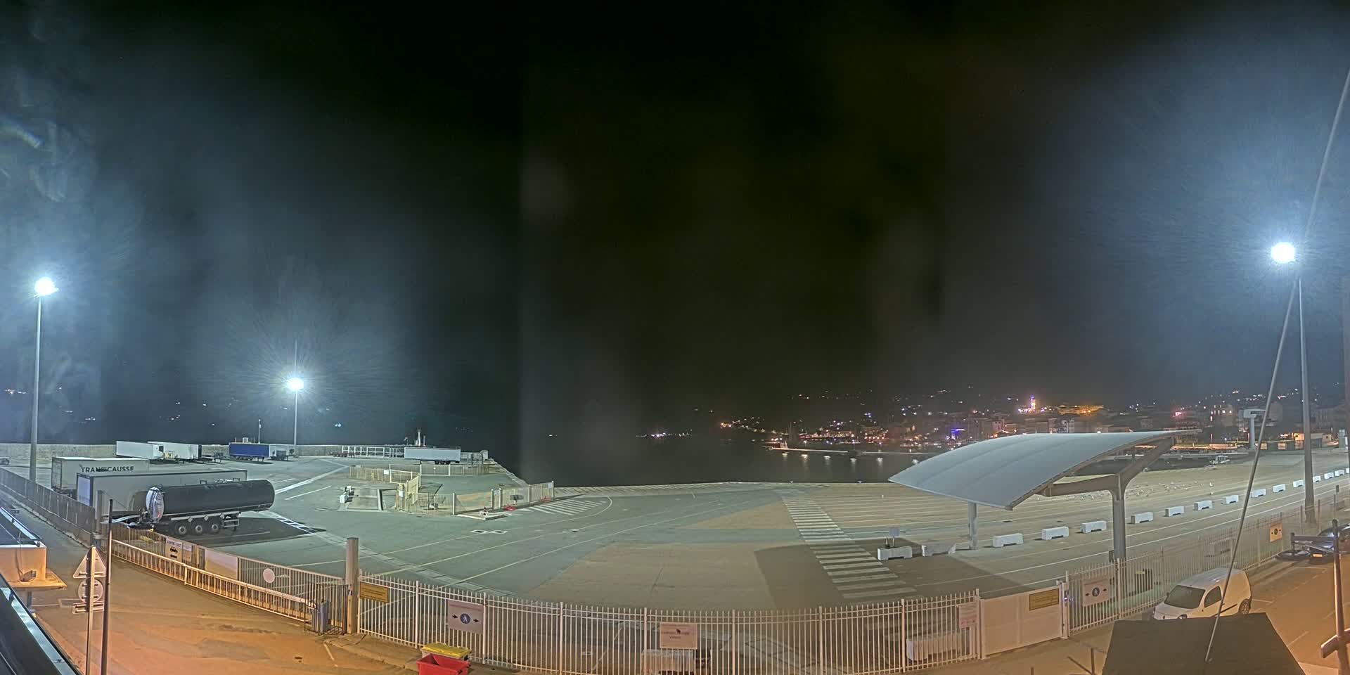 A nighttime view of a mostly empty port area with a tanker truck and a few other vehicles, under a dark, slightly hazy sky.