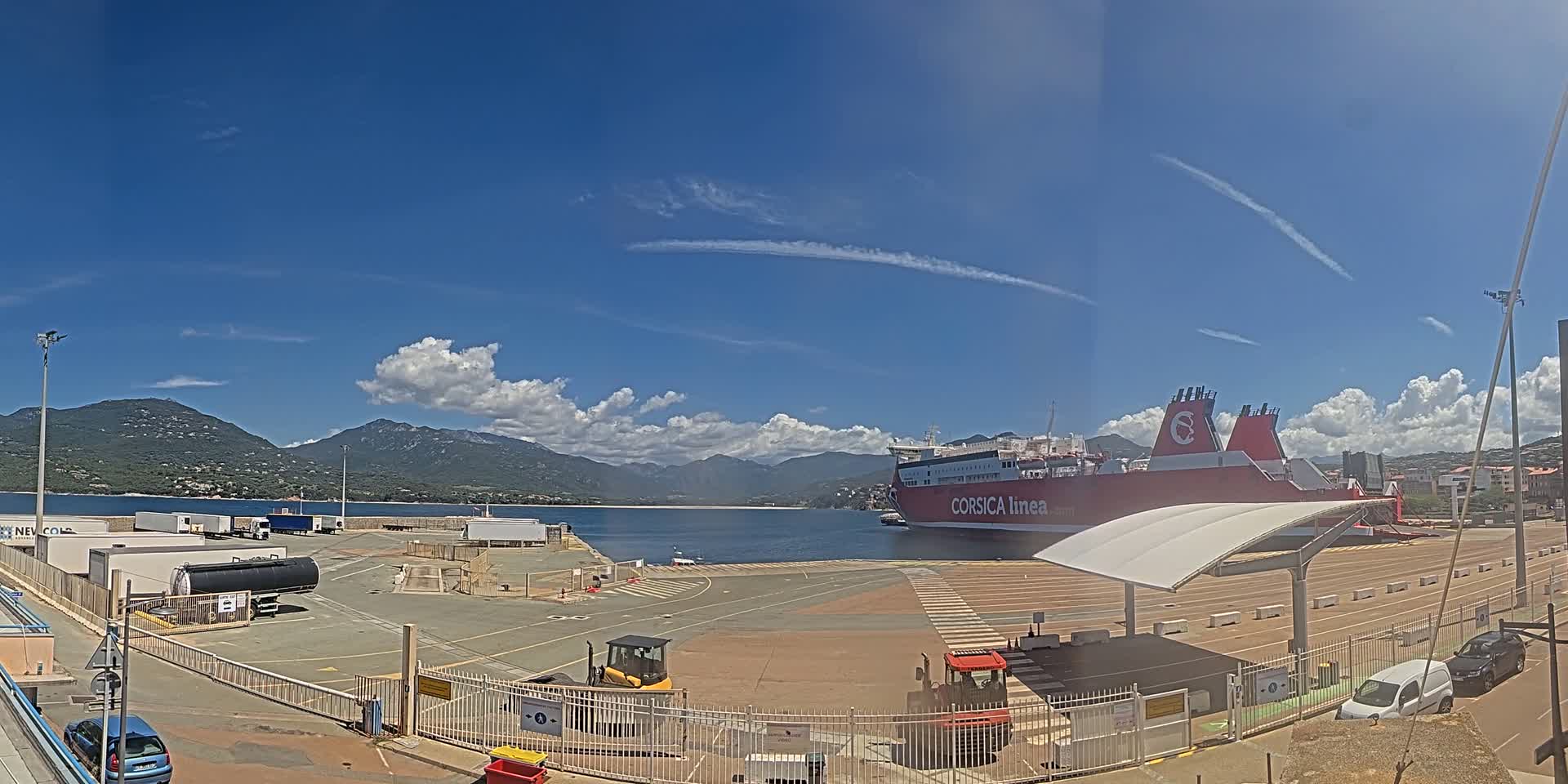 A large red ferry is docked at a port under a mostly sunny sky with scattered clouds and mountains in the background.
