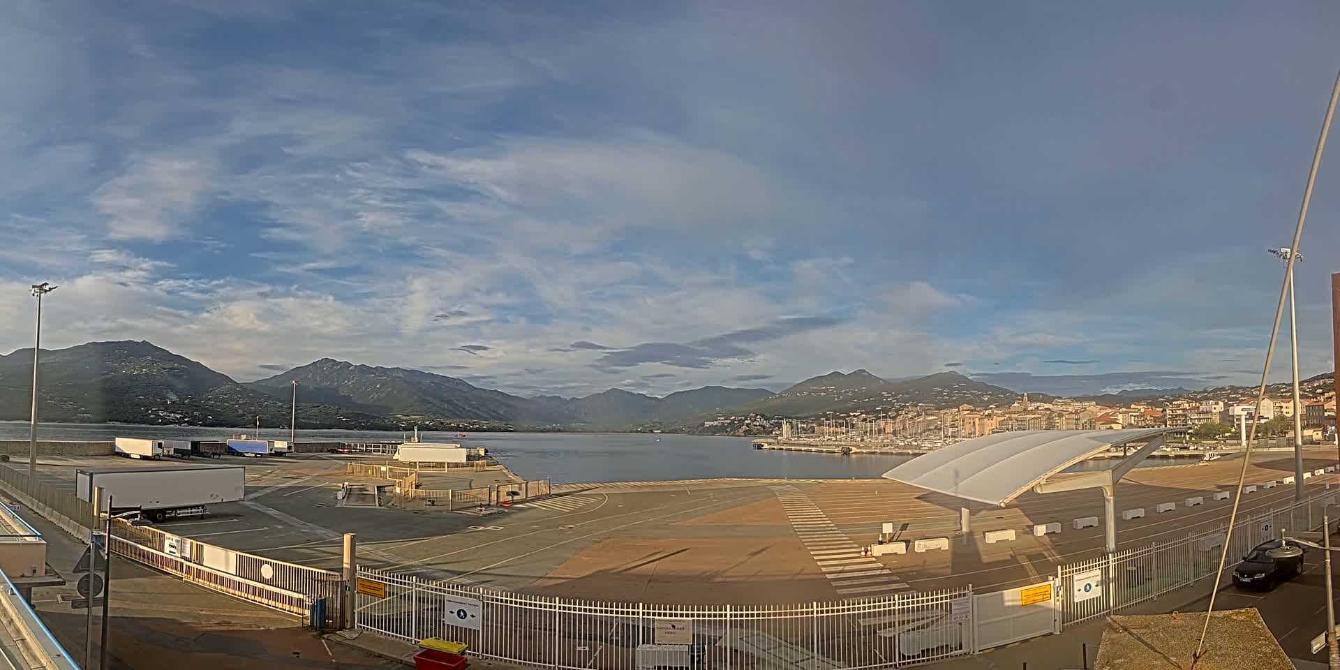 A mostly sunny day reveals a harbor with several docked boats, a large empty paved area, and mountains in the background.