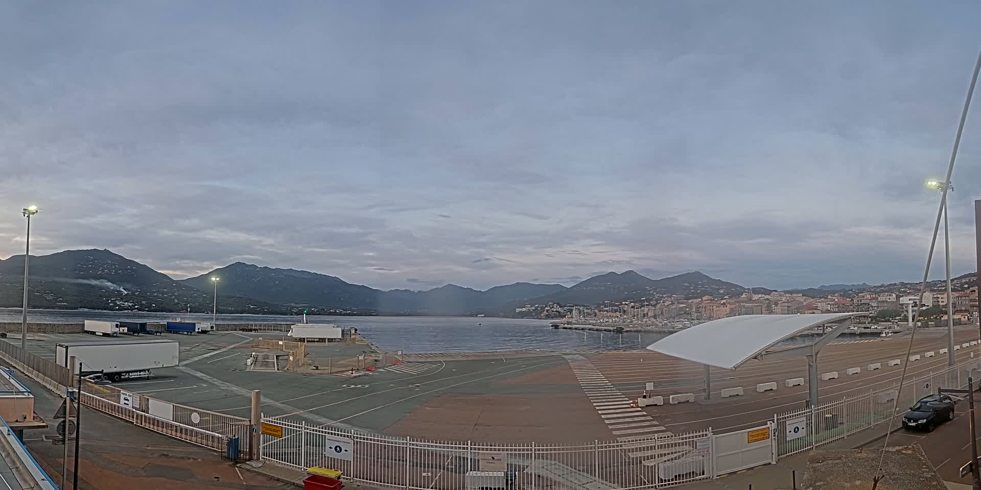 An empty port area with several trucks and a car, under a cloudy sky, overlooks a calm body of water and mountains in the distance.