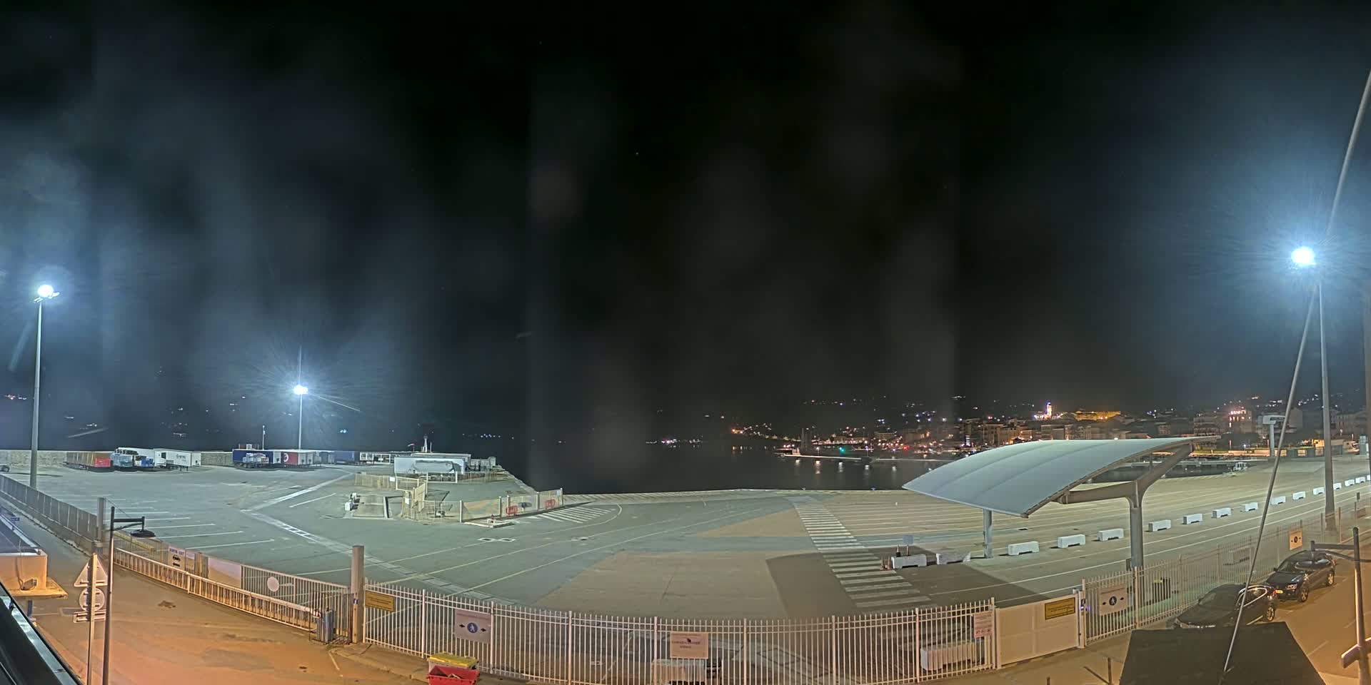 A nighttime view of an empty port area with several trucks parked, brightly lit by floodlights, and a town visible in the distance under a hazy, dark sky.