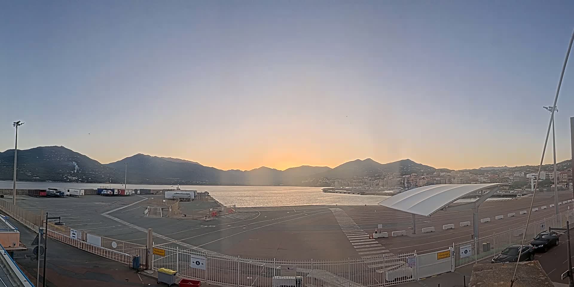 A mostly empty port area with a few vehicles, bordered by a metal fence, overlooks a calm body of water and mountains at sunset under a clear sky.