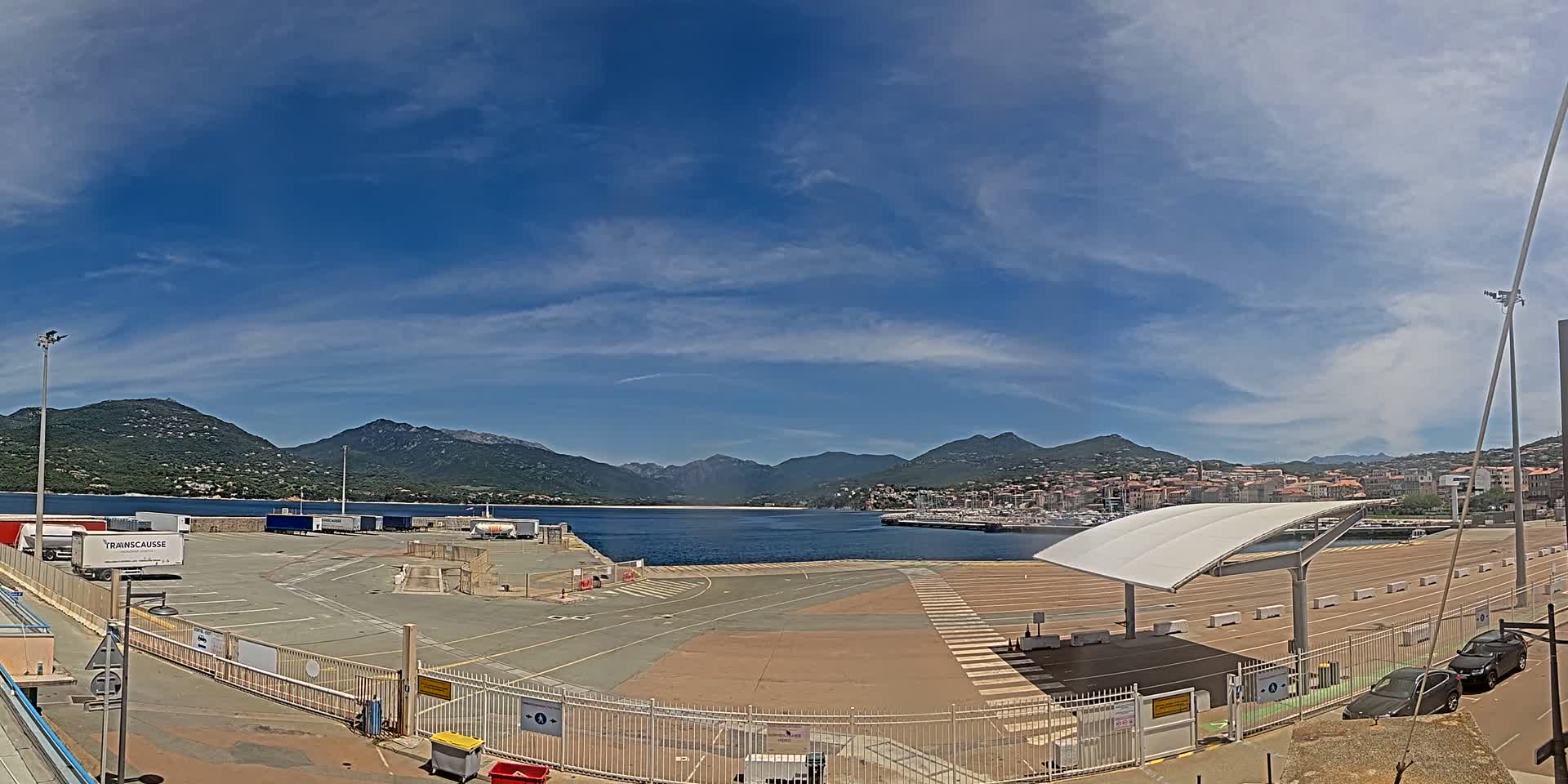 A mostly sunny day reveals a port area with a large empty paved lot, several parked trucks, a marina with boats, and mountains in the background.