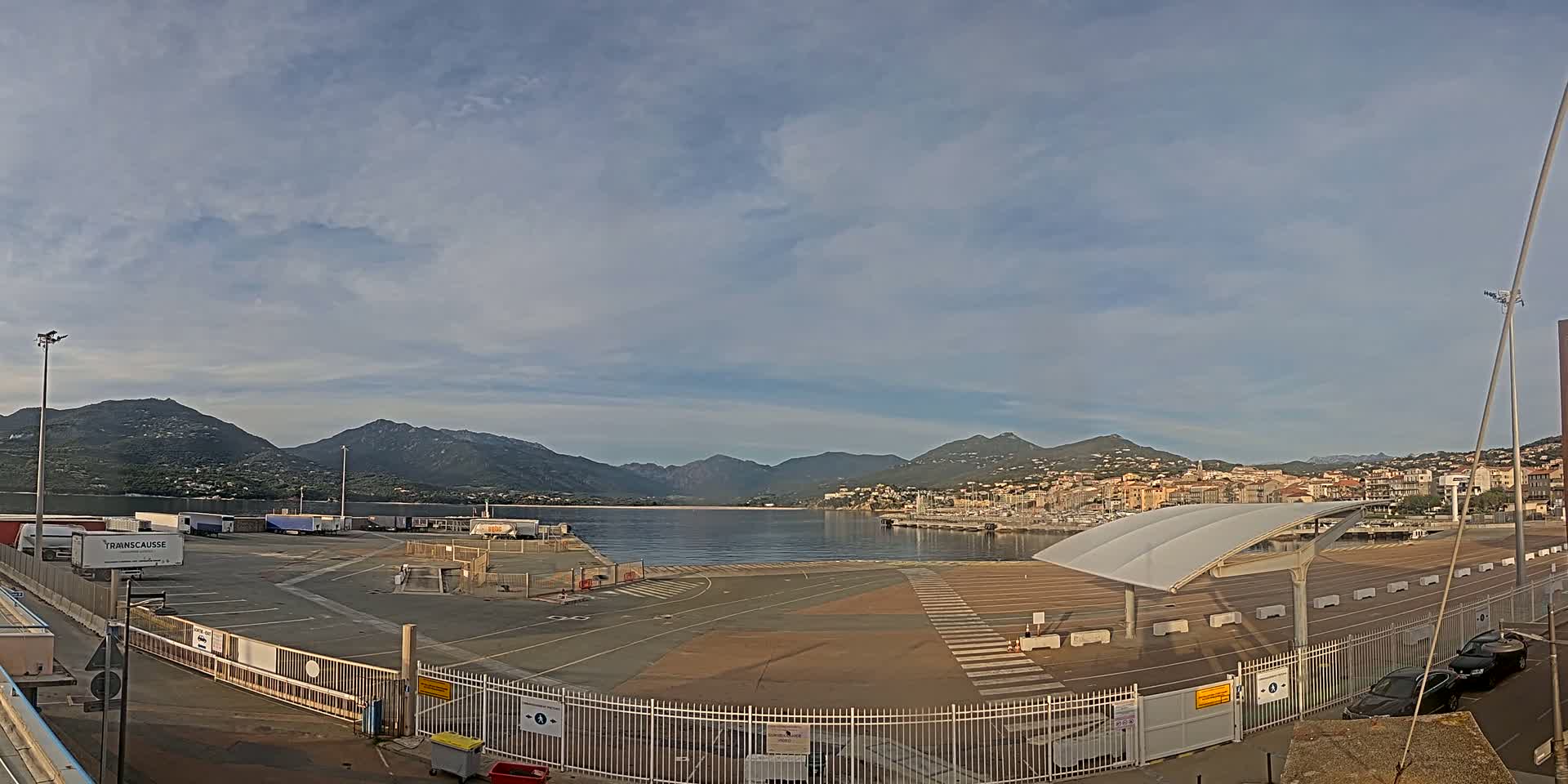 An empty port area with a few parked cars and trucks, a covered waiting area, and mountains in the background under a partly cloudy sky.