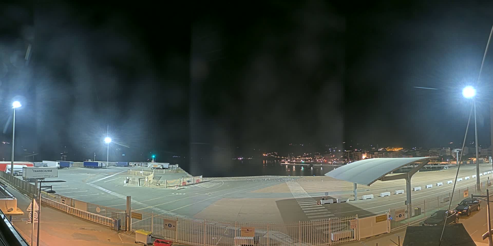 A nighttime view of an empty port area with several parked trucks and a cityscape in the distance under hazy conditions.