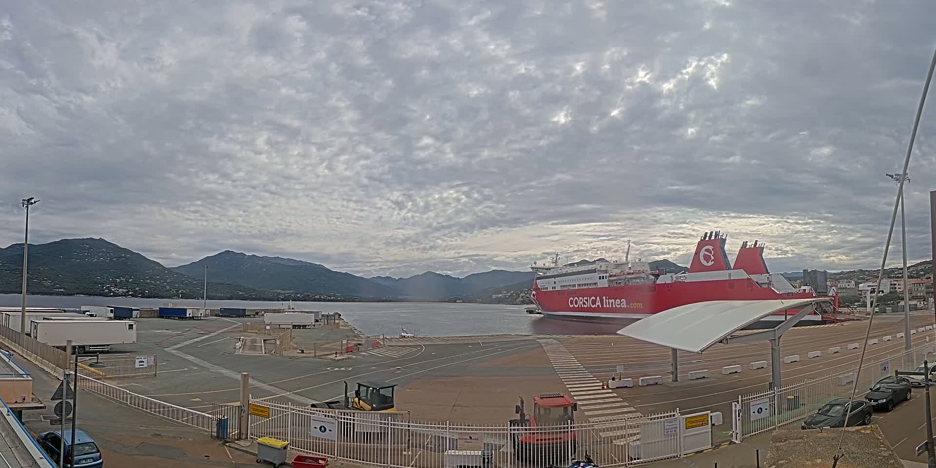 A large red ferry is docked at a port, under a cloudy sky, with mountains in the background.