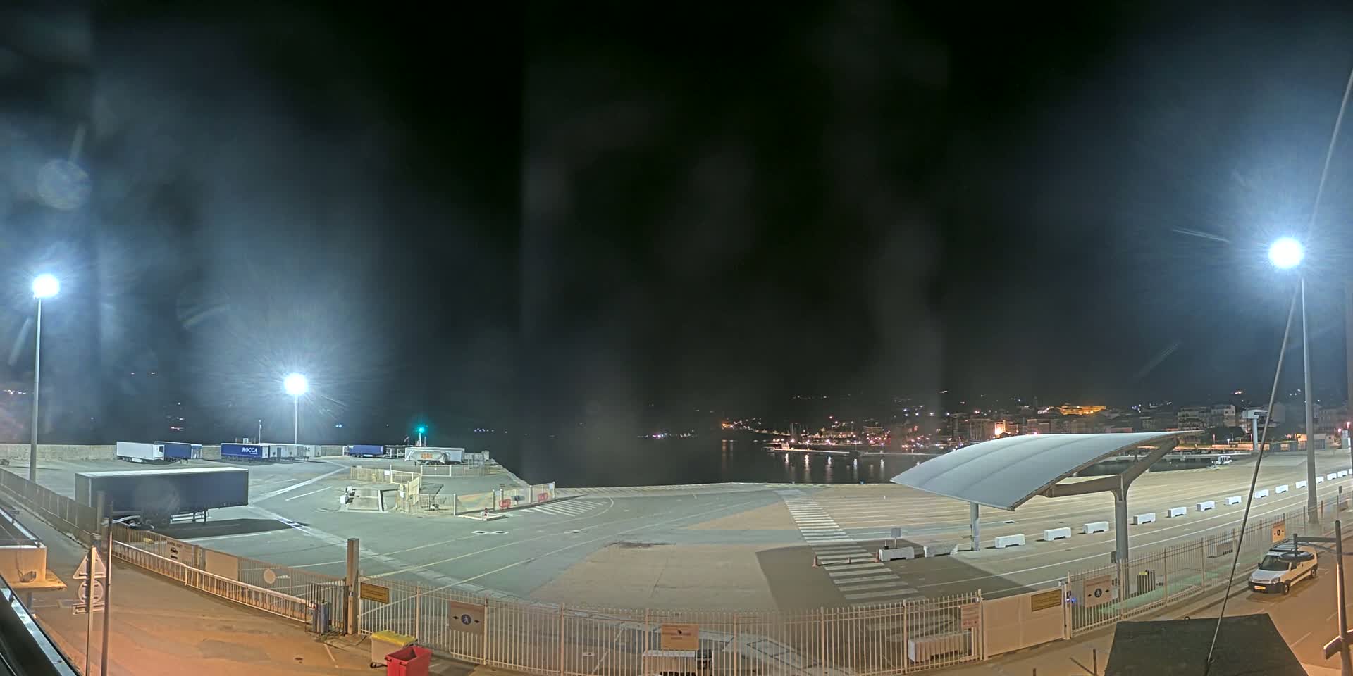 A nighttime view of a port area with several trucks parked, a covered waiting area, and a town visible in the distance under a hazy sky.