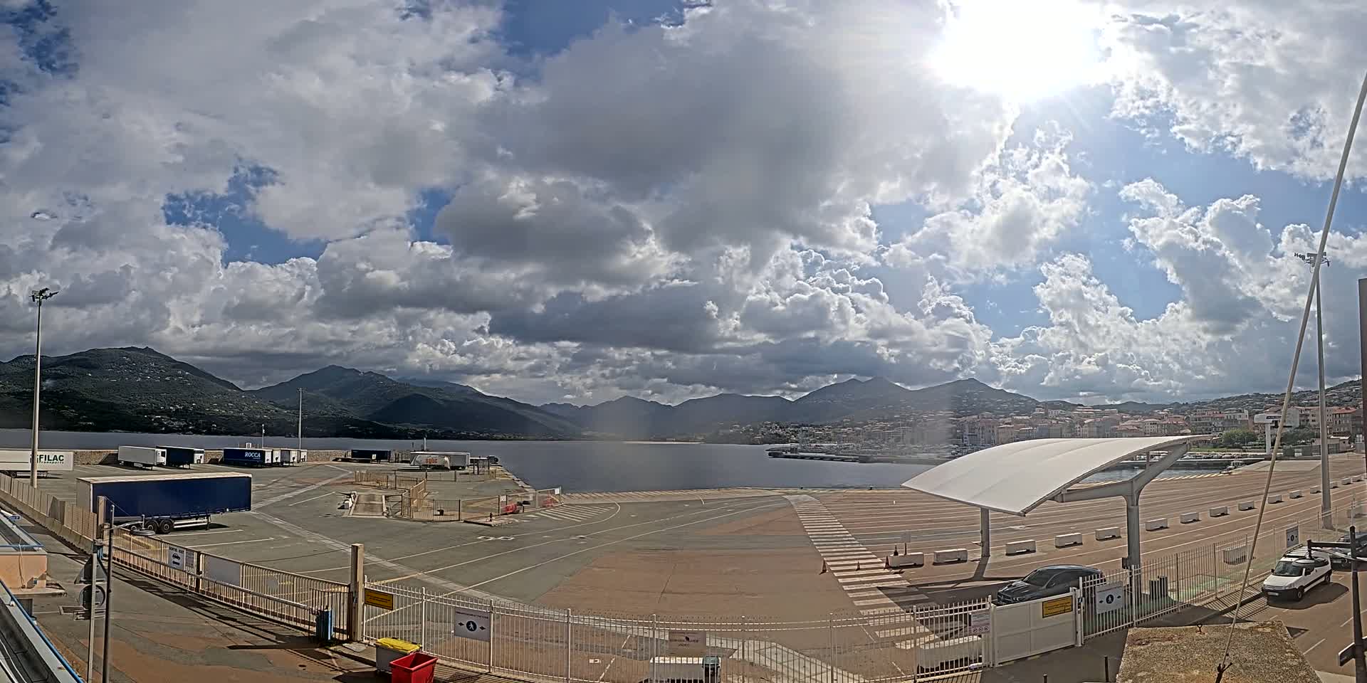 A partly sunny day reveals a harbor with several trucks parked in a lot next to a body of water backed by mountains.