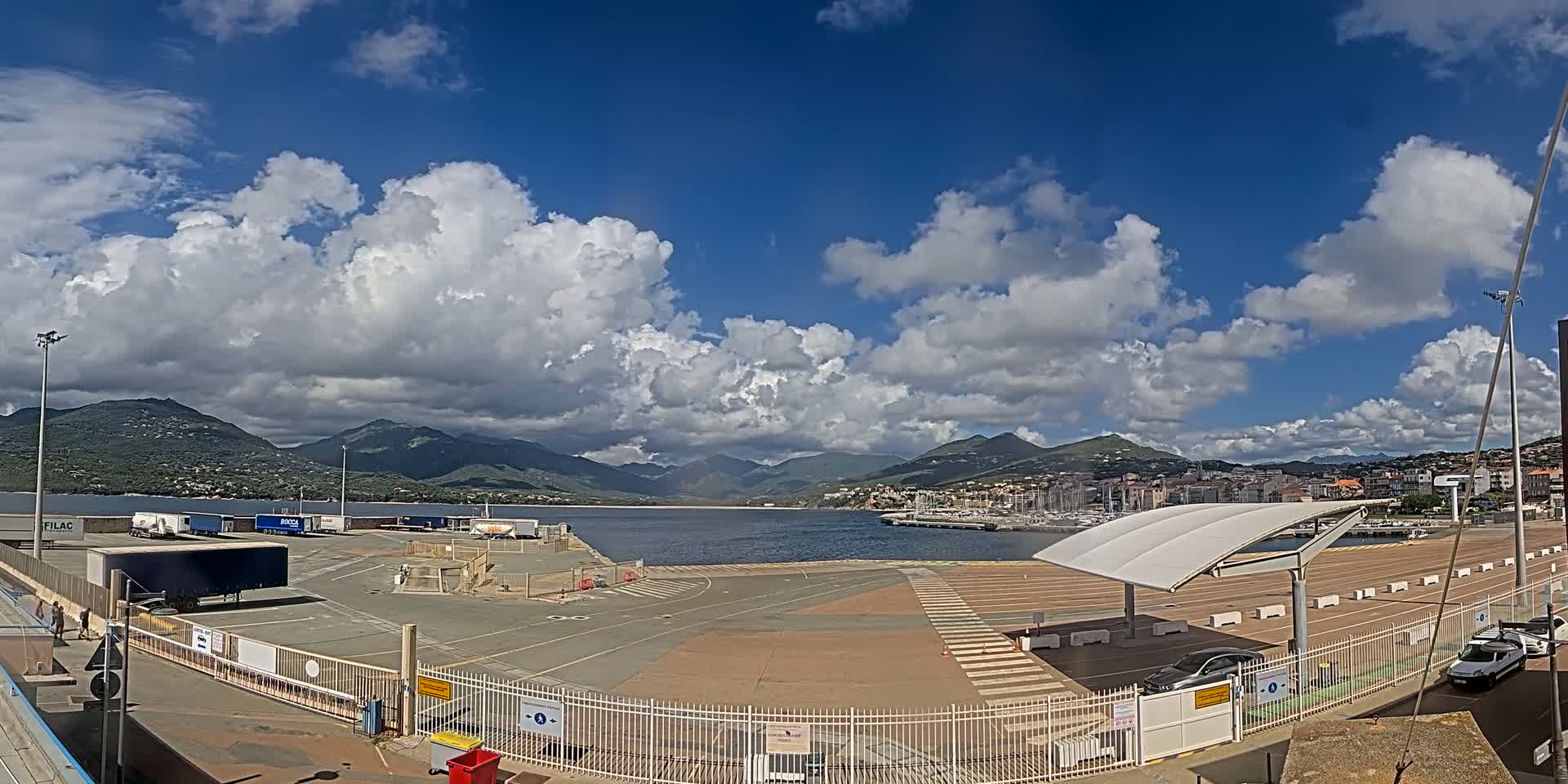 A partly cloudy sky over a harbor with mountains in the background, featuring a large paved area with several trucks and cars, and a covered waiting area.