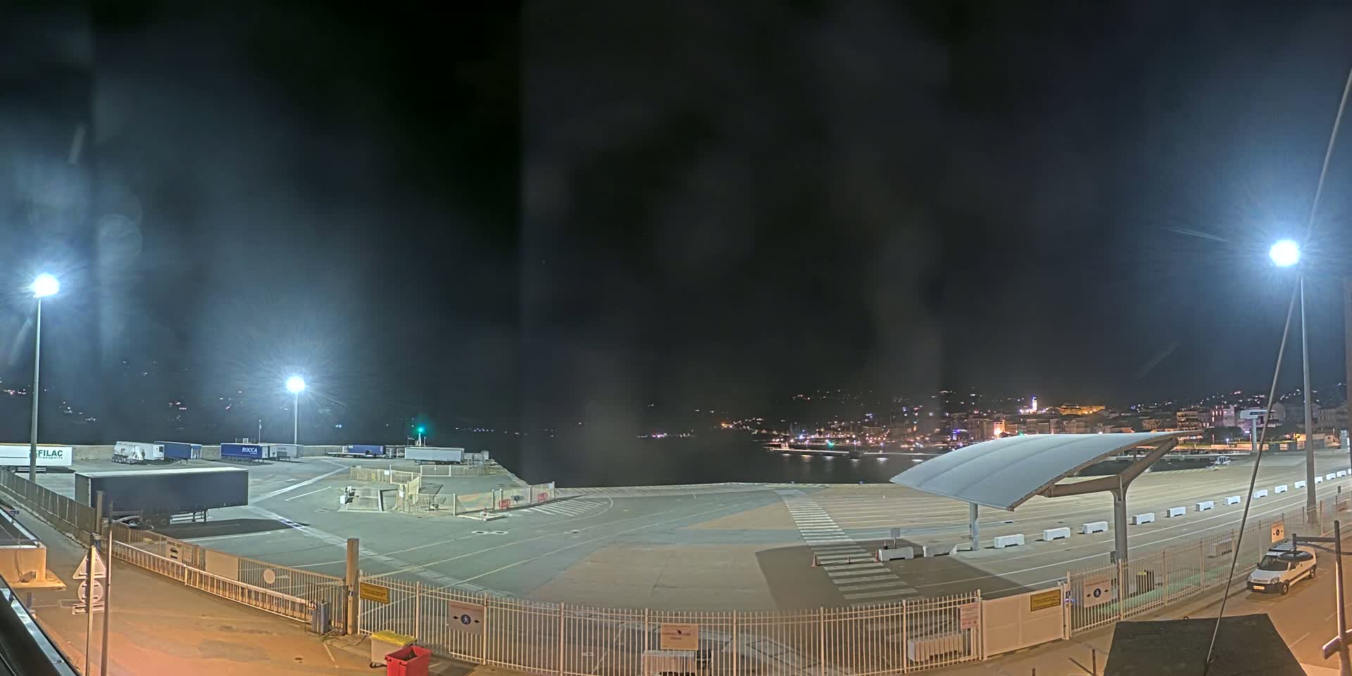 A nighttime view of a mostly empty port area with several parked semi-trailer trucks and a distant, illuminated cityscape across the water.