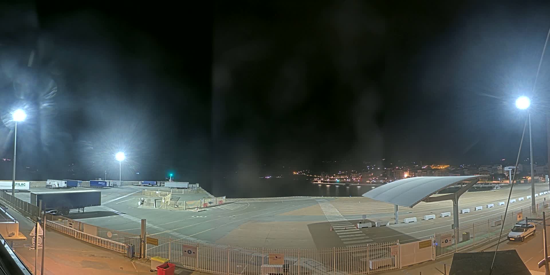 A nighttime view of a port area with several trucks, a covered waiting area, and a town visible in the distance under hazy, night sky conditions.