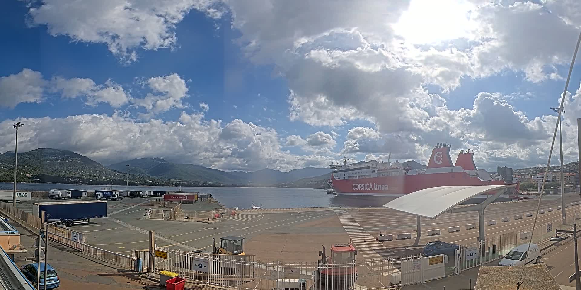 A large red ferry is docked at a port under a partly cloudy sky, with mountains visible in the background.