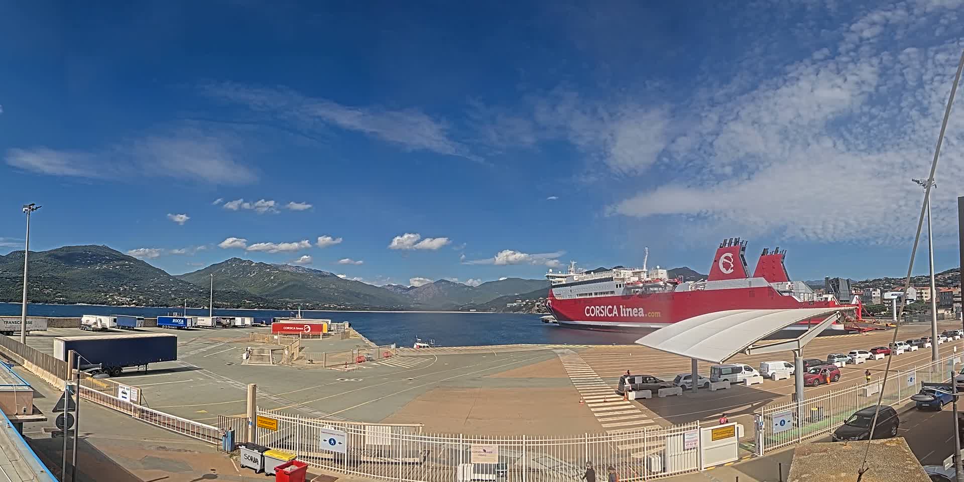 A large red ferry is docked at a port under a mostly sunny sky, with mountains in the background and several vehicles parked nearby.