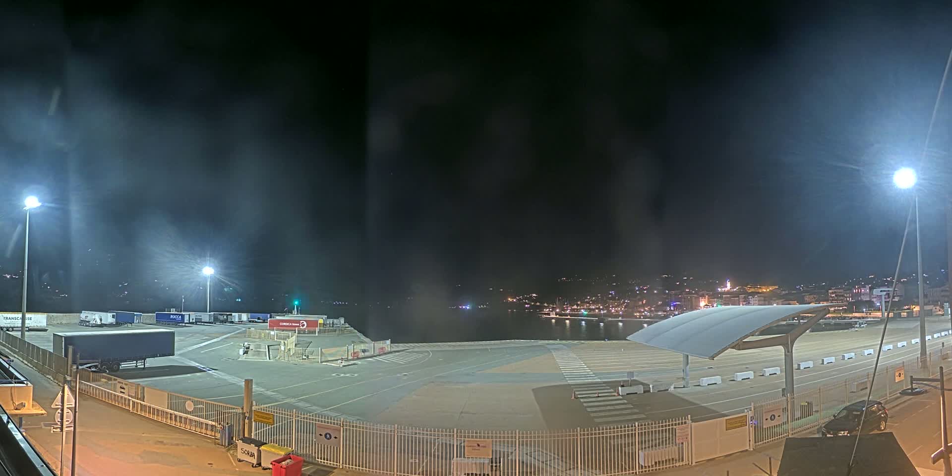 A nighttime view of a port area with several parked trucks and a covered waiting area, overlooking a calm body of water with a city skyline in the distance under a dark, slightly hazy sky.