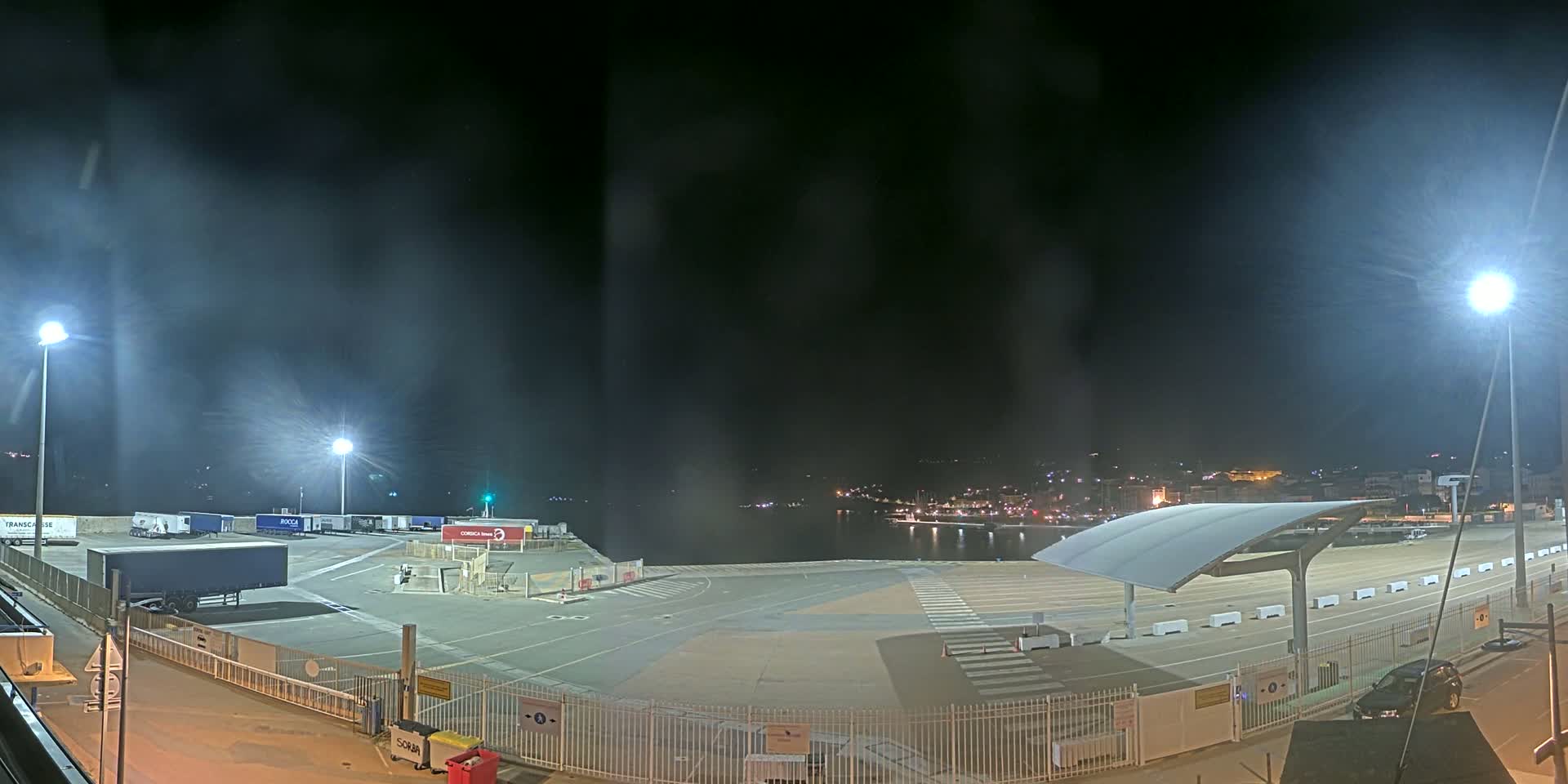 A nighttime view of a port area with several parked trucks and a covered waiting area, under hazy conditions.