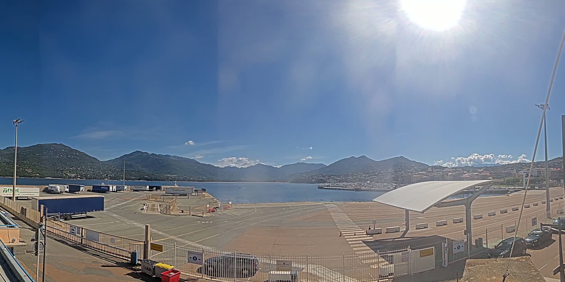 A sunny day at a harbor with several parked trucks, cars, and a modern canopy structure, overlooking a calm body of water and mountains in the distance.