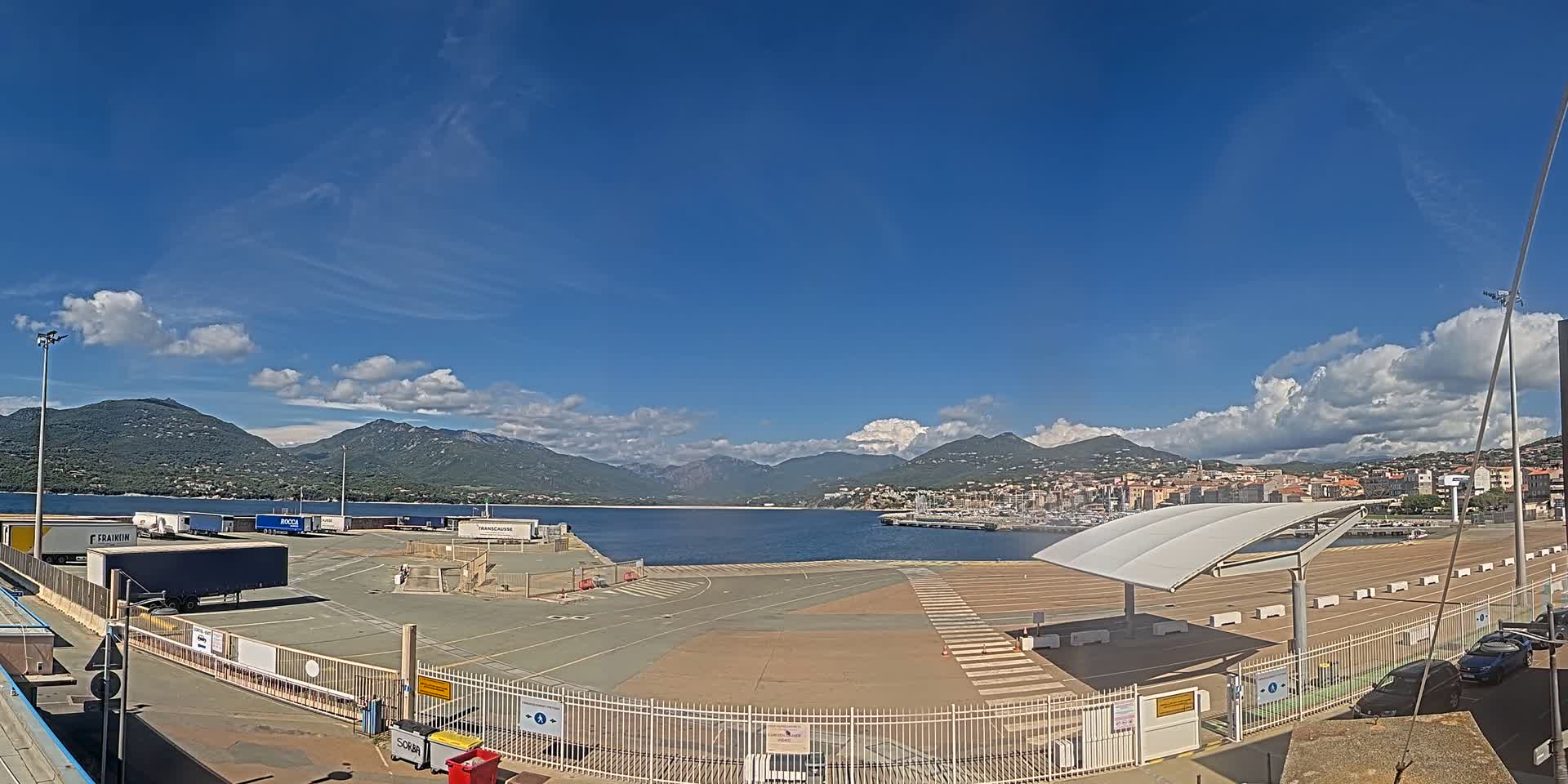 A mostly sunny day reveals a port area with several trucks and a marina, mountains in the background, and a few cars parked near a gated entrance.