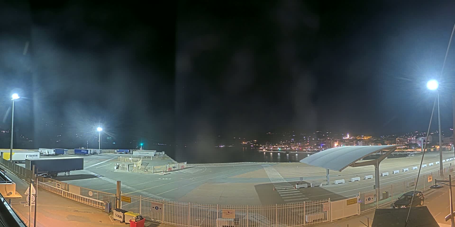 An empty, brightly lit port area at night, with several parked trucks and a distant city skyline visible across the water.