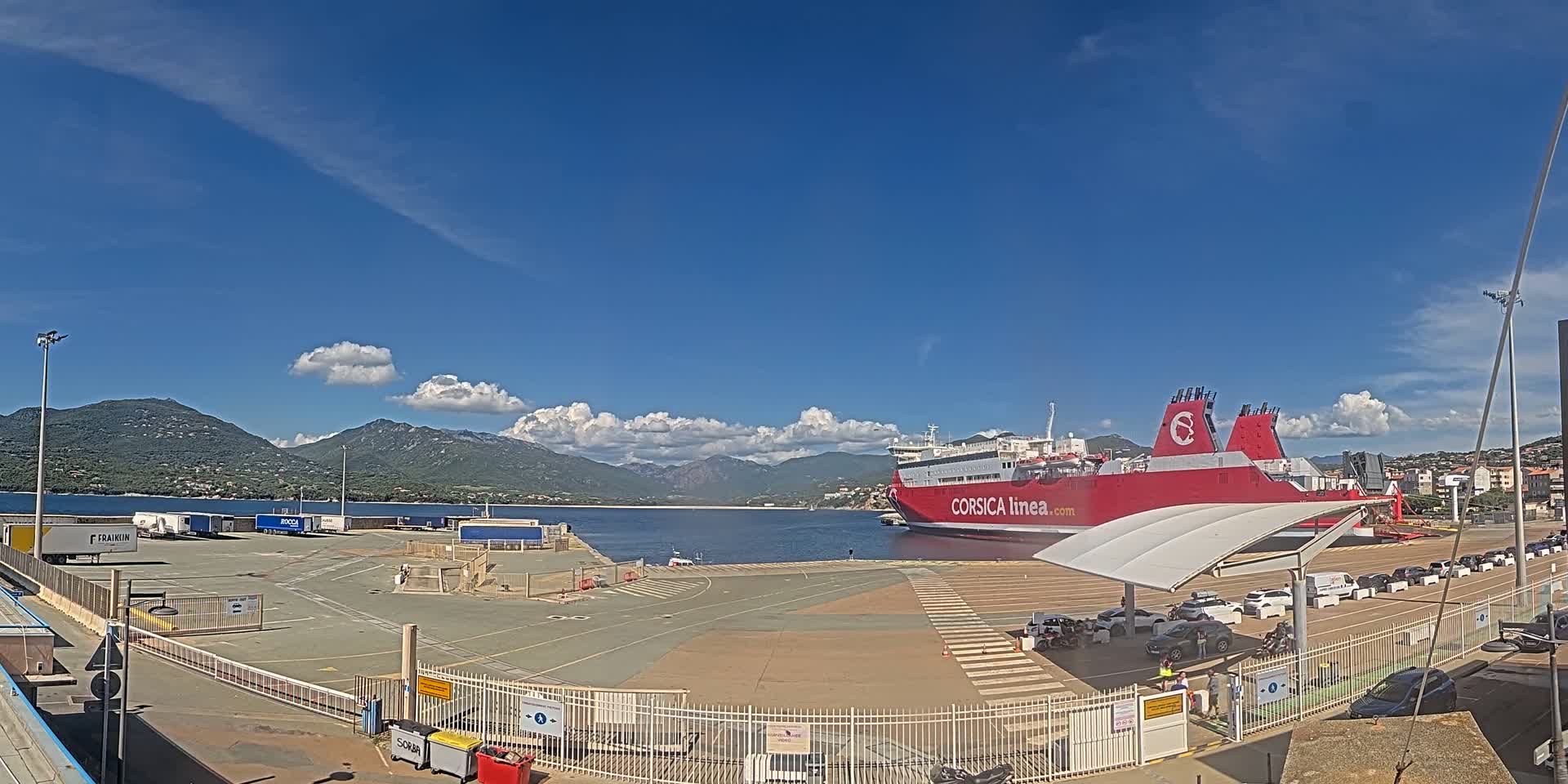 A large red ferry is docked at a port under a mostly sunny sky with some fluffy clouds, mountains are visible in the distance.