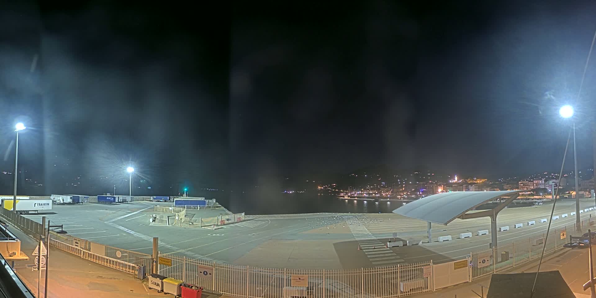 An empty, brightly lit nighttime port area with several parked trucks and a distant town visible across the water.
