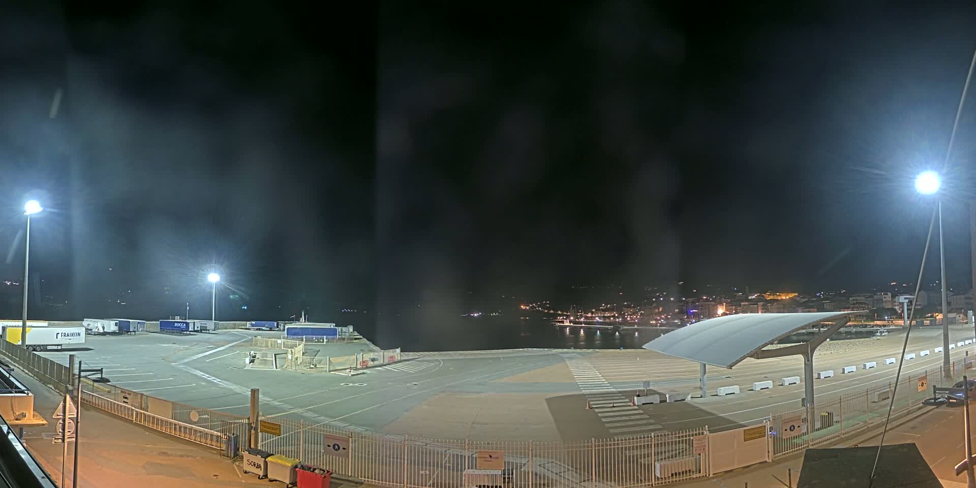A nighttime view of an empty port area with several parked trucks, illuminated by bright lights, overlooking a calm body of water and a distant town.