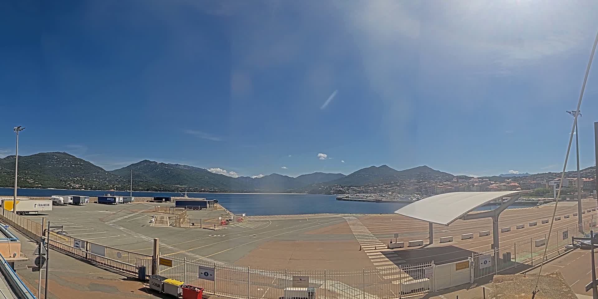 A sunny day at a port with a large empty paved area, several cargo containers, and mountains in the background.