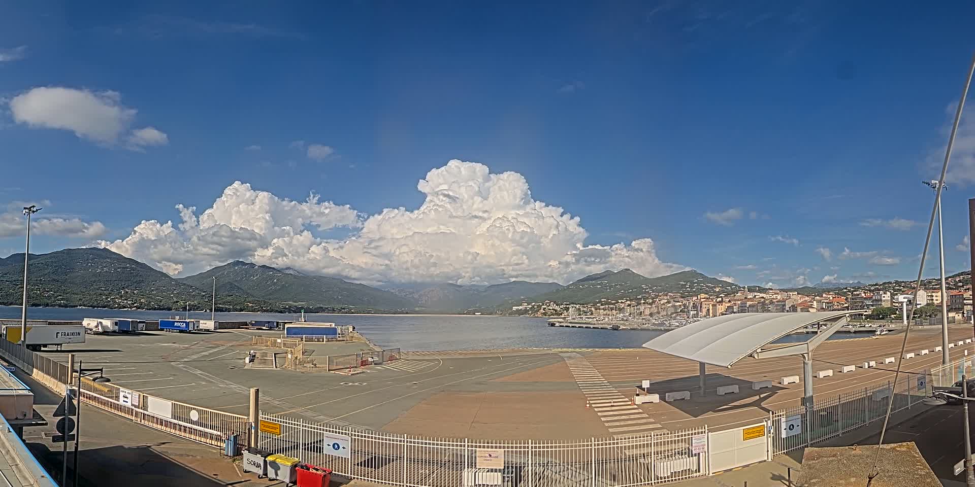 A port with numerous semi-trucks and a marina is visible under a partly cloudy sky, with mountains in the background.