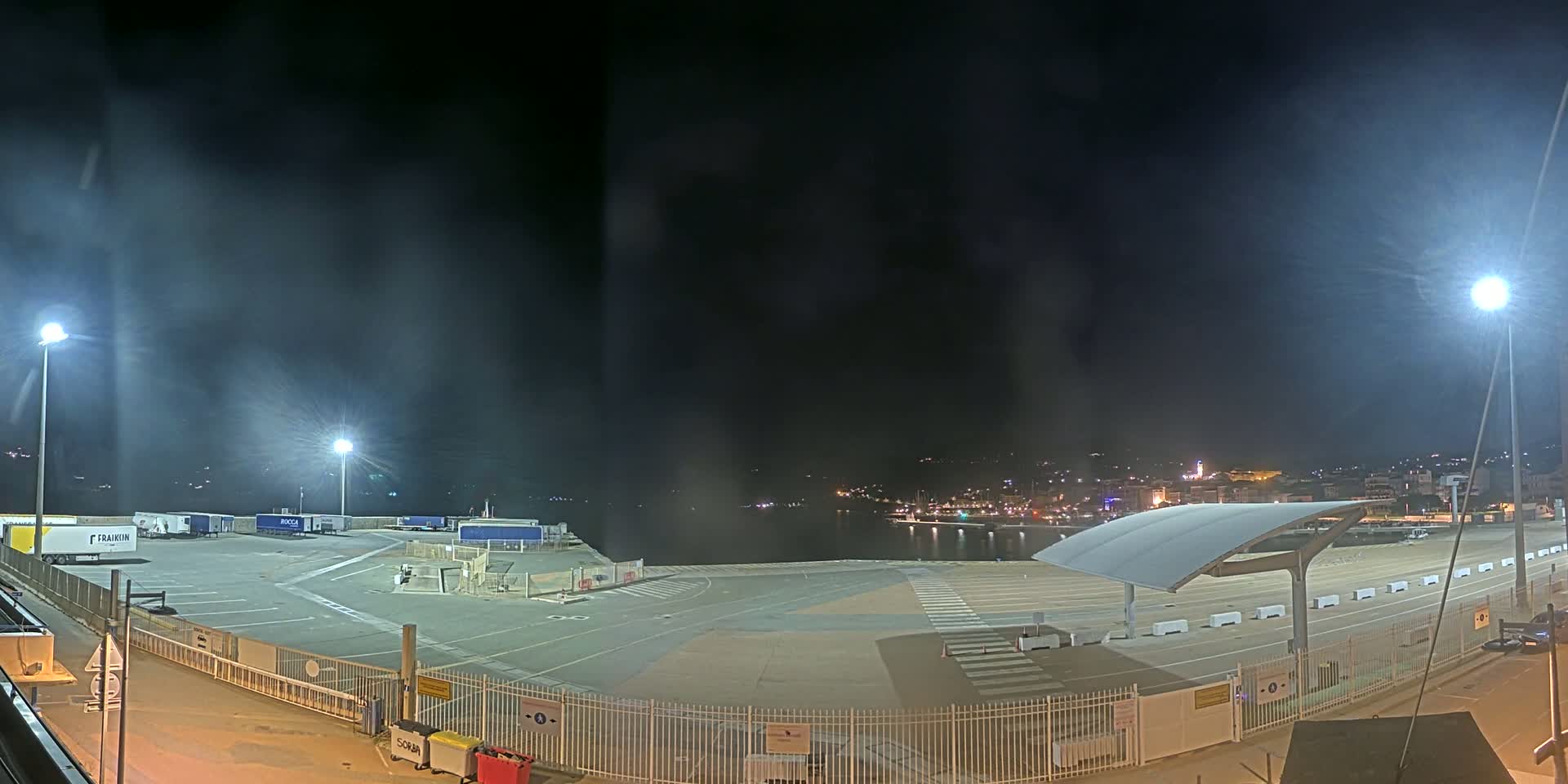 A nighttime view of a port area with several trucks parked, a mostly empty paved lot, and a town visible across the water under a slightly hazy sky.