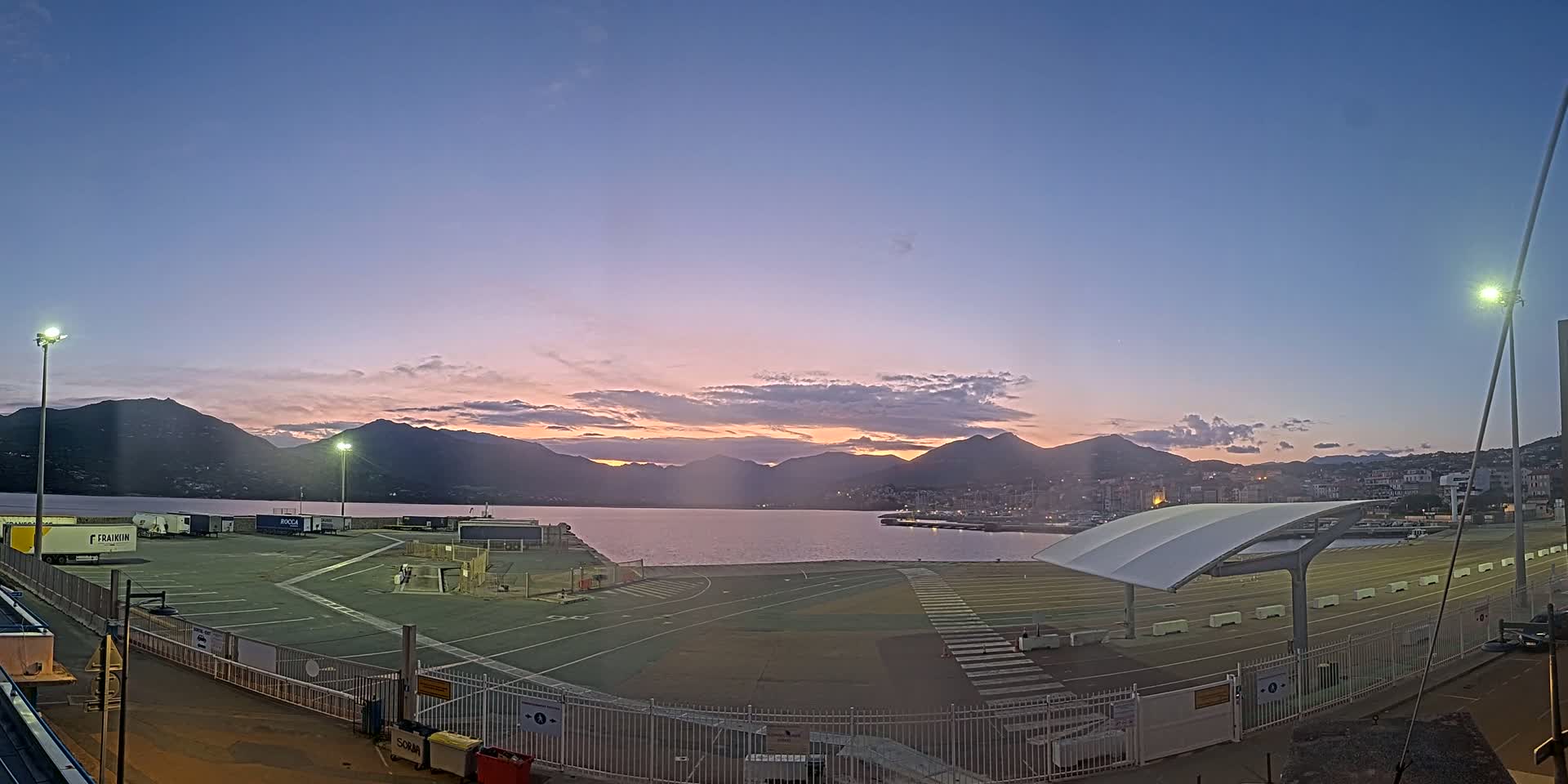A calm body of water is seen at sunset, behind a paved area with parked trucks and a covered waiting area, all bordered by mountains in the background.