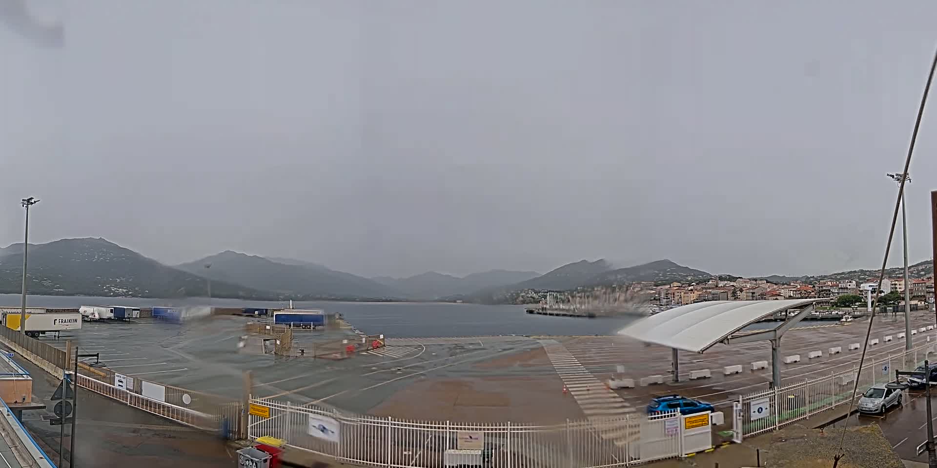 A wet, empty parking lot and ferry terminal overlook a calm body of water and distant mountains under an overcast sky.