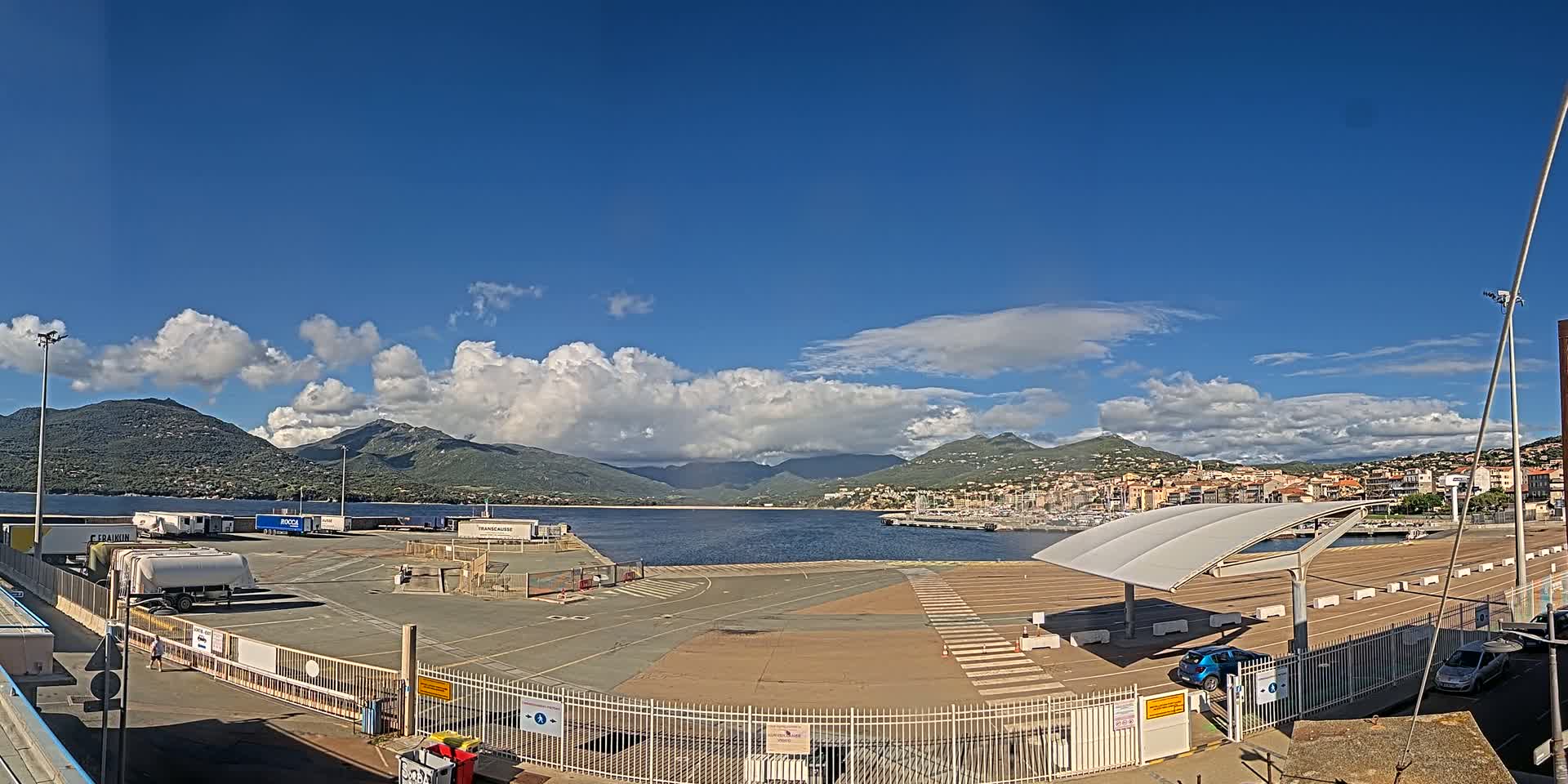 A port area with a large paved lot, some parked vehicles, and a few buildings is situated near a calm body of water, with mountains in the background under a partly cloudy, sunny sky.