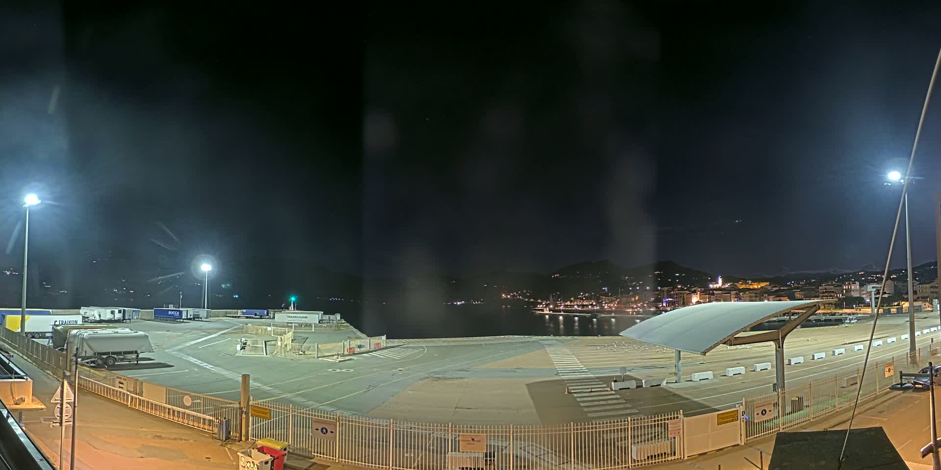 A nighttime view of an empty port area with several parked trucks and trailers, illuminated by bright lights, overlooking a calm body of water and a distant town nestled against hills.
