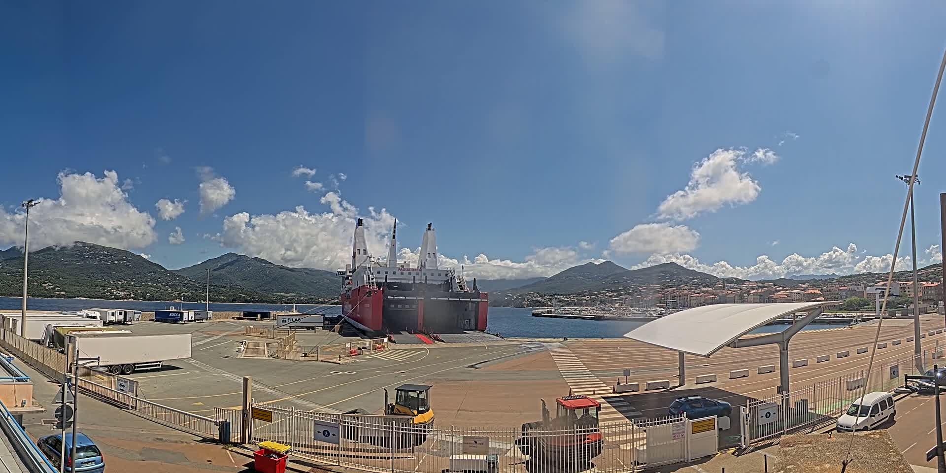 A large red and white ferry is docked at a port, under a mostly sunny sky with some fluffy clouds, and mountains are visible in the distance.