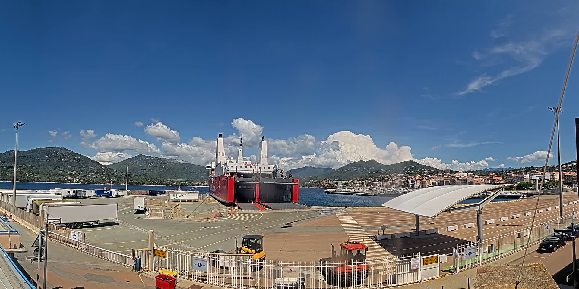A large red ferry is docked at a port on a sunny day with partly cloudy skies and mountains in the background.