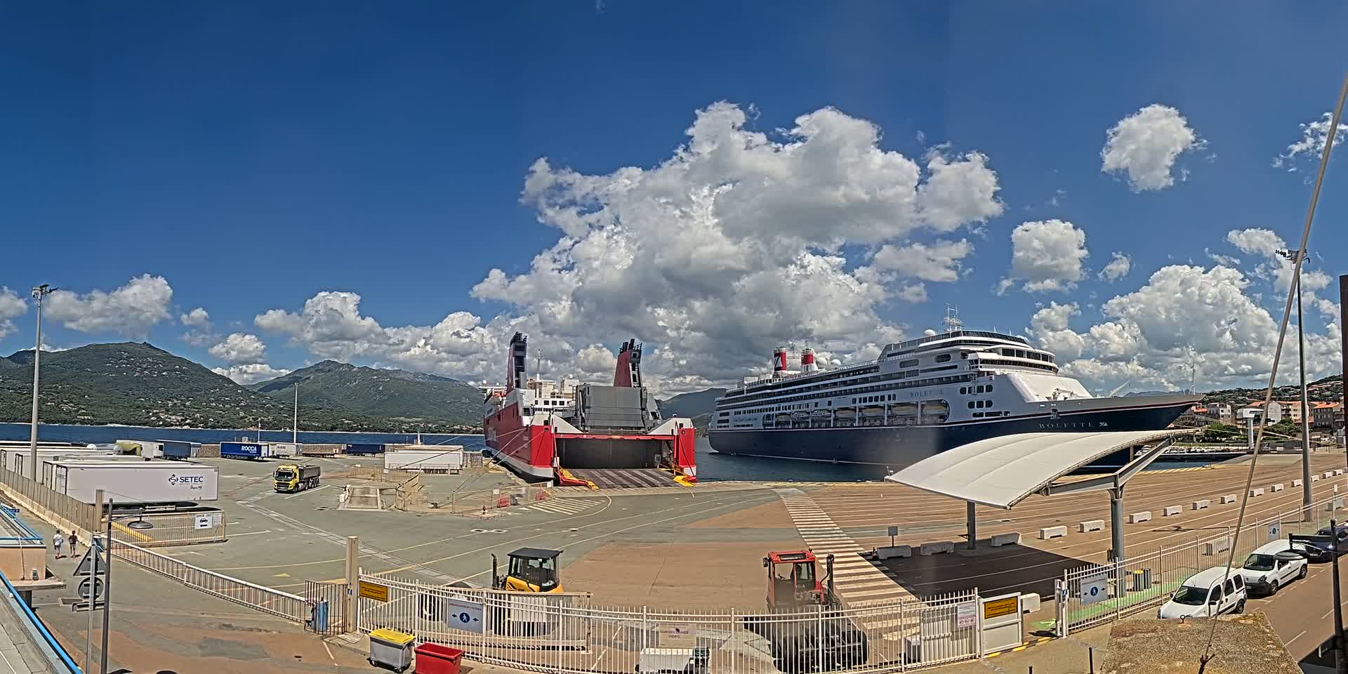 A ferry and a cruise ship are docked at a port under a partly cloudy sky, with mountains visible in the background.