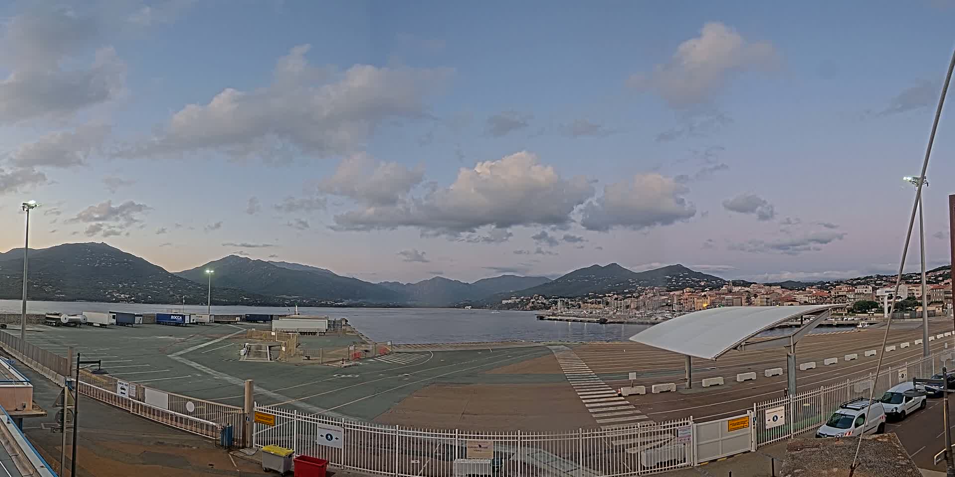 A mostly empty port with a few vehicles, under a partly cloudy sky, overlooks a calm body of water and mountains in the distance.