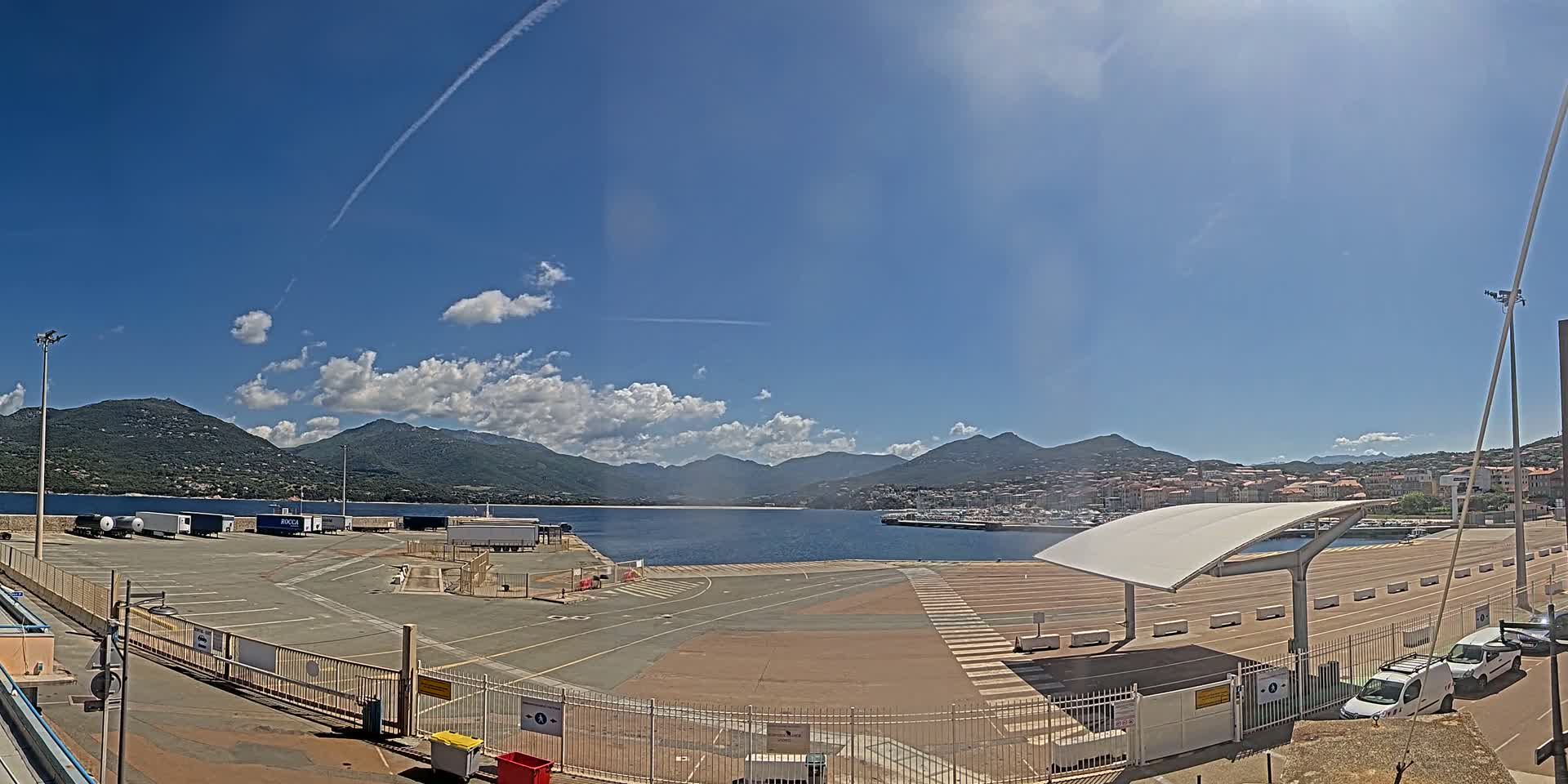 A sunny day at a port with mountains in the background, featuring a large paved area with parked trailers and cars, and a covered waiting area.